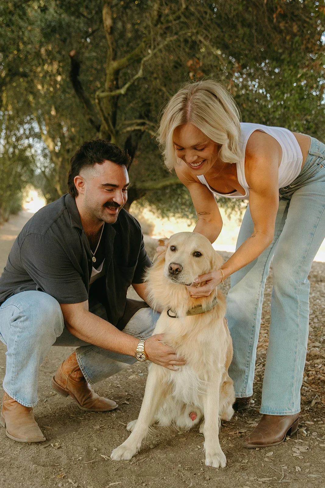Playful moment of the couple kneeling together petting their golden retriever under soft evening light.