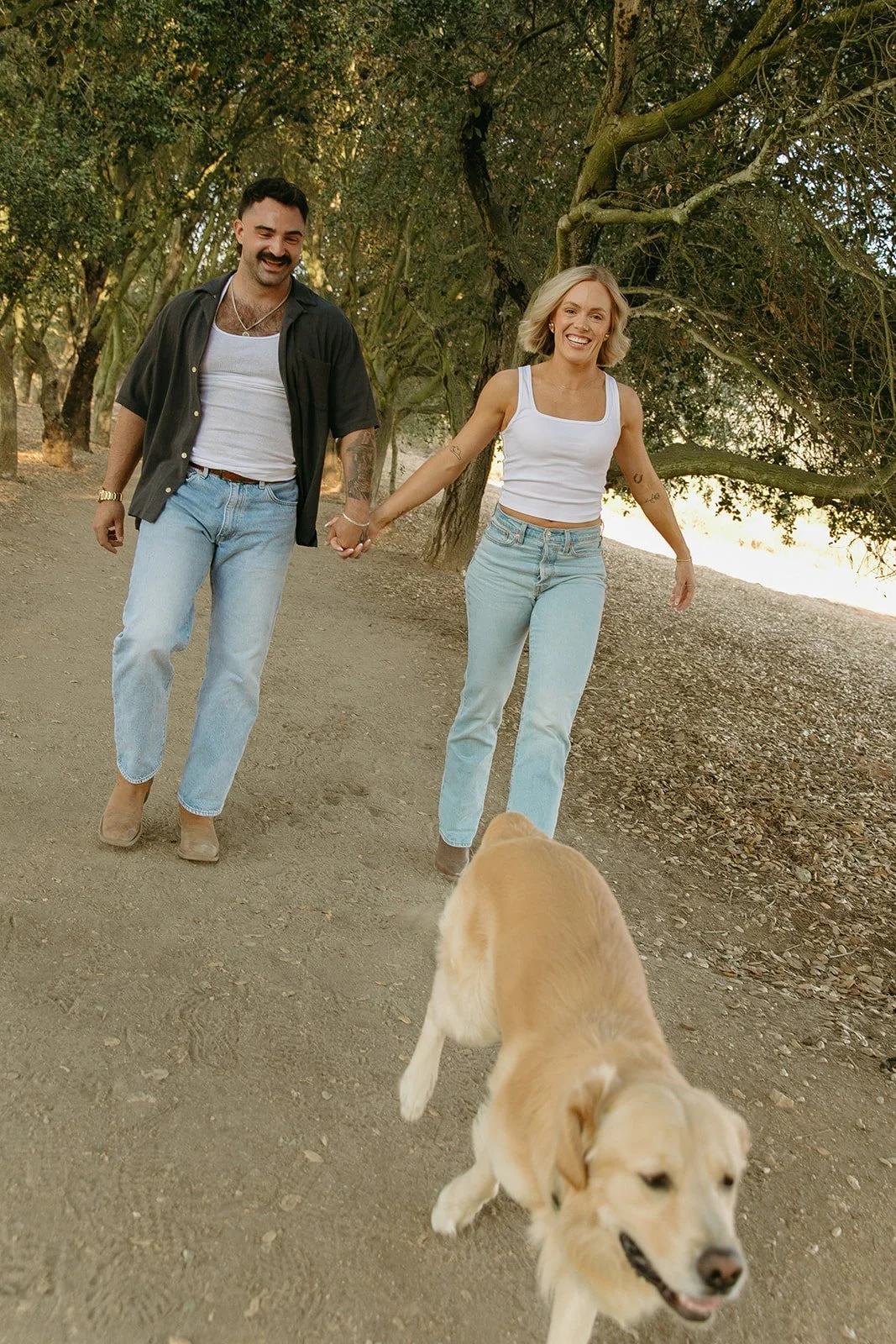 Candid western engagement photos of the couple running hand in hand down a wooded trail with their dog leading the way.