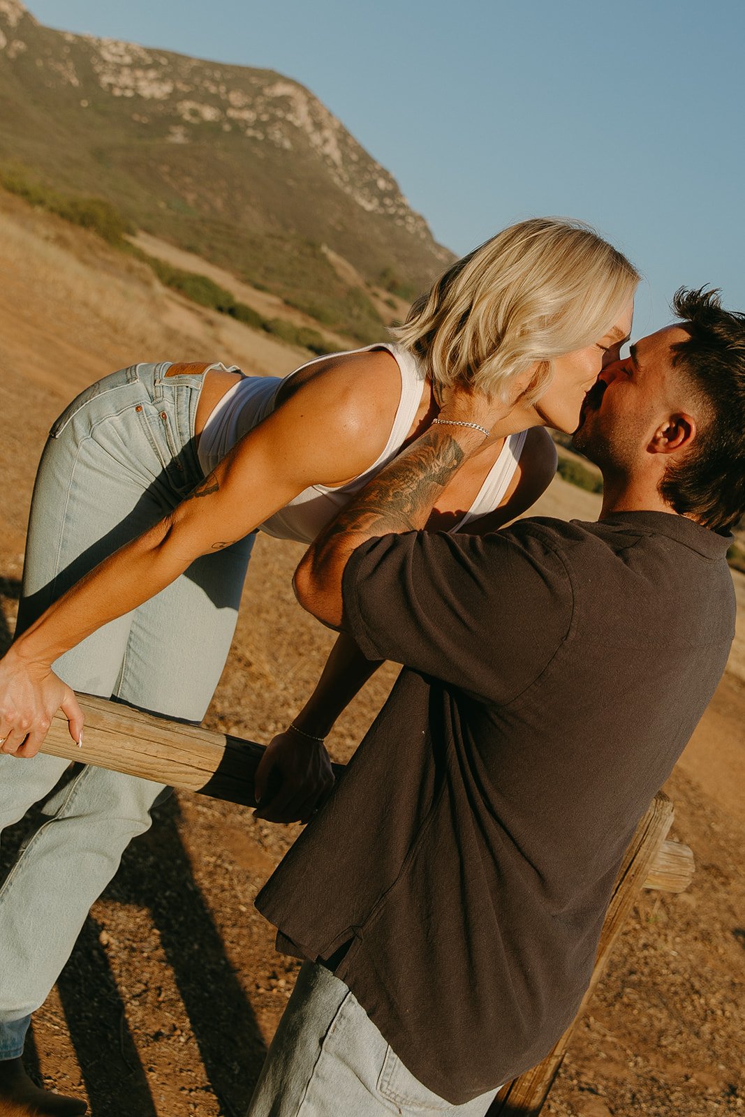 Romantic kiss captured in warm sunset light, showcasing laid-back western engagement photos with denim outfits and rustic fencing.