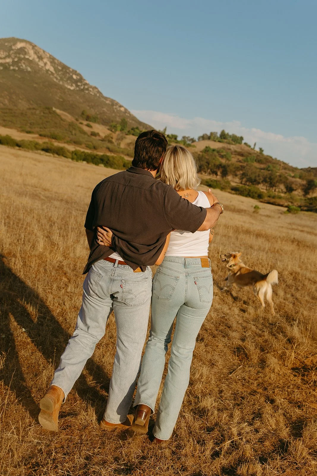 Scenic western engagement photos of the couple walking through an open field at sunset with their dog running beside them.