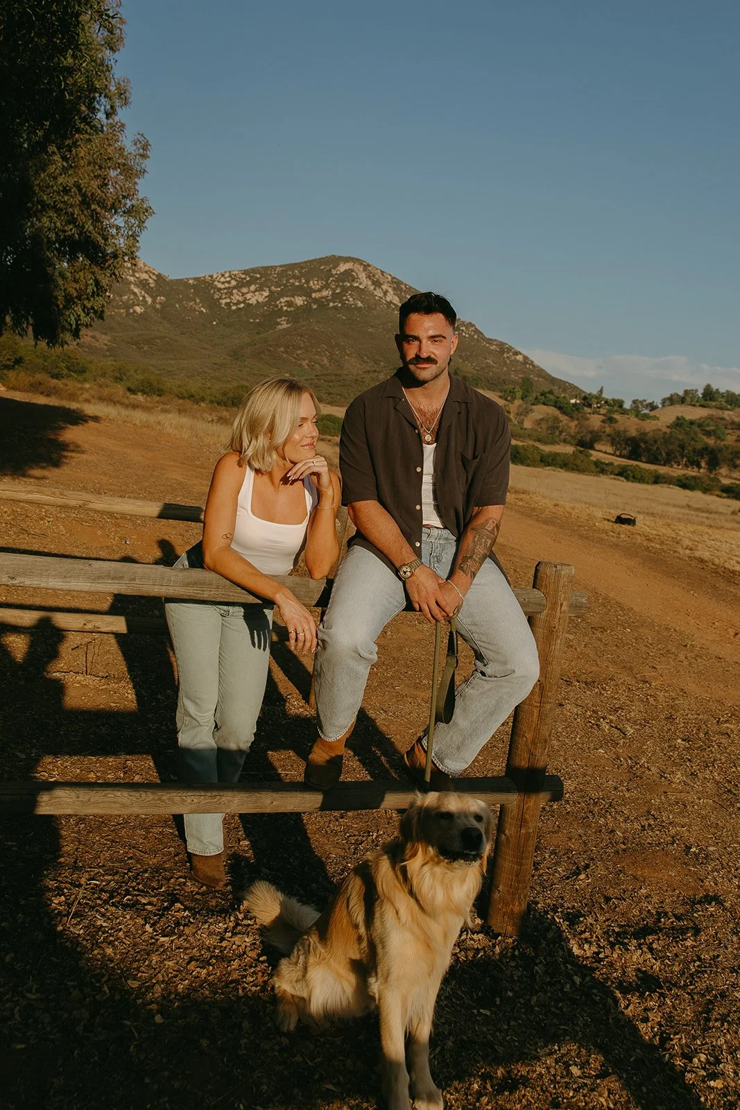 Candid portrait of the couple sitting on a fence together with their dog below them, mountains stretching across the background.