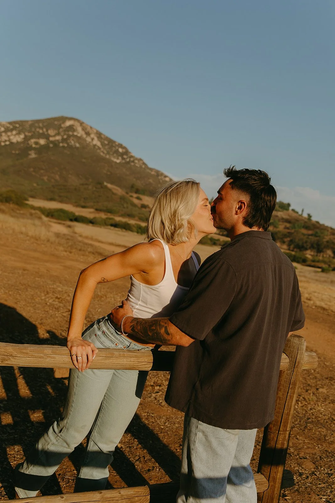 Wide candid moment of the couple leaning over a fence sharing a kiss in the middle of a sunlit mountain landscape.