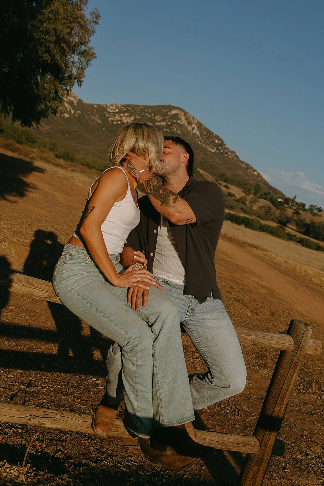 Golden hour western engagement photos of the couple sitting on a wooden fence kissing with rolling hills and mountains behind them.