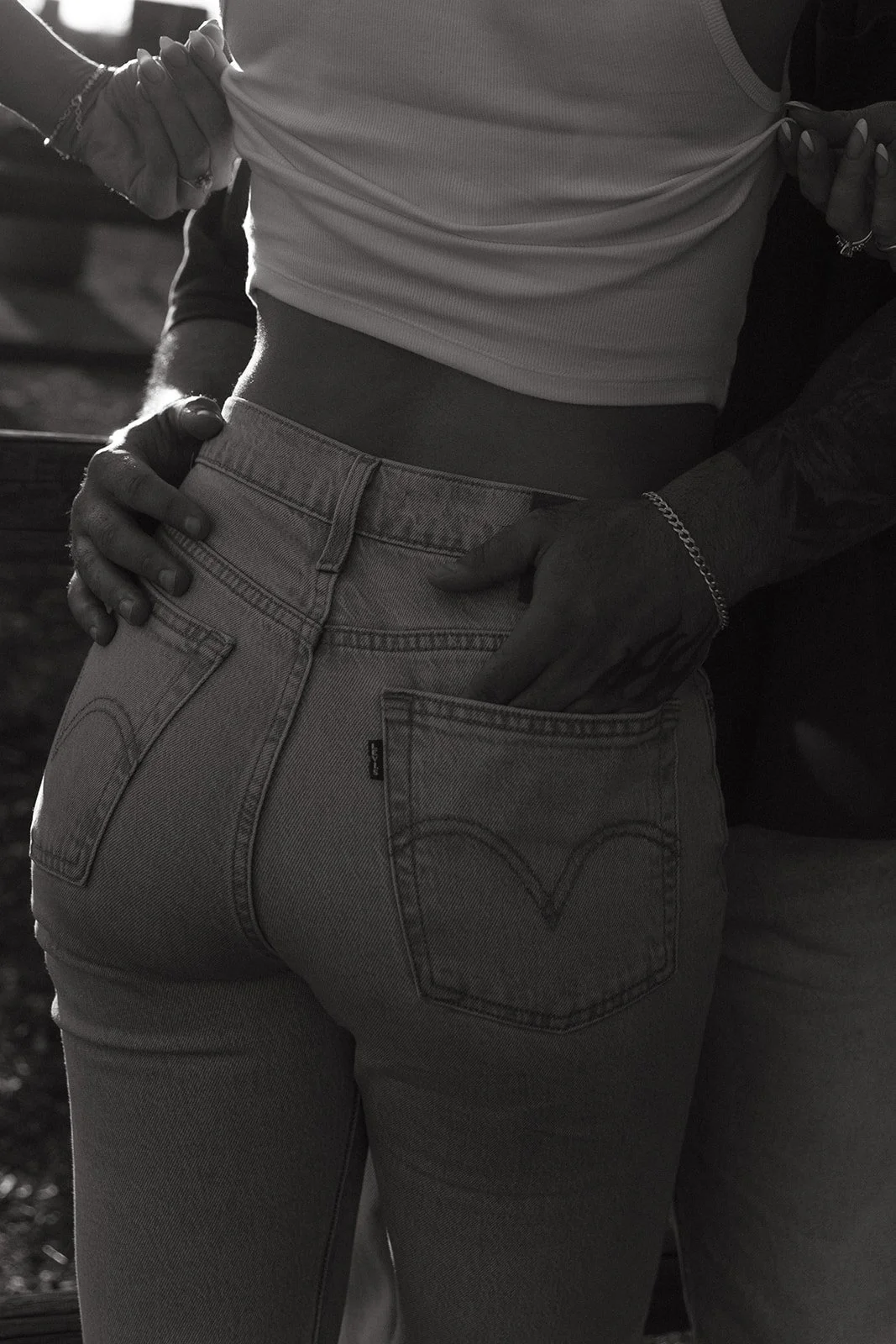Black and white close up of the couple standing together, hands wrapped around her waist as she lifts her tank slightly, showcasing an intimate, candid moment.