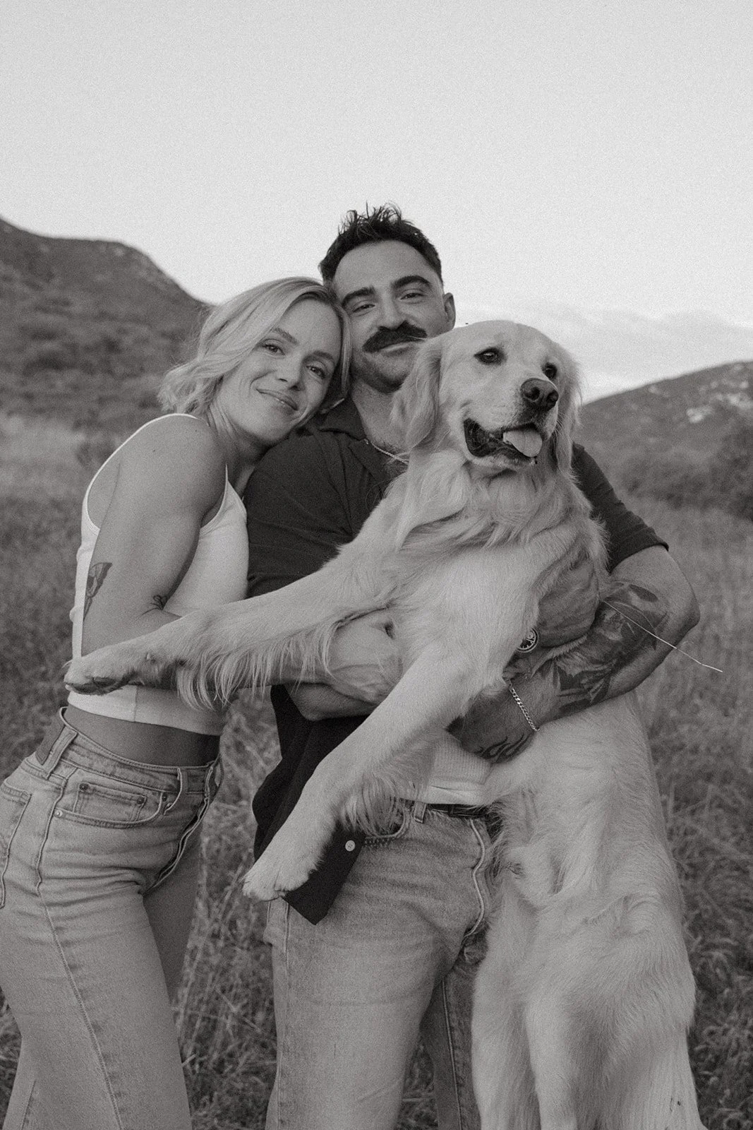 Black and white portrait of the couple smiling while holding their golden retriever together in an open meadow.