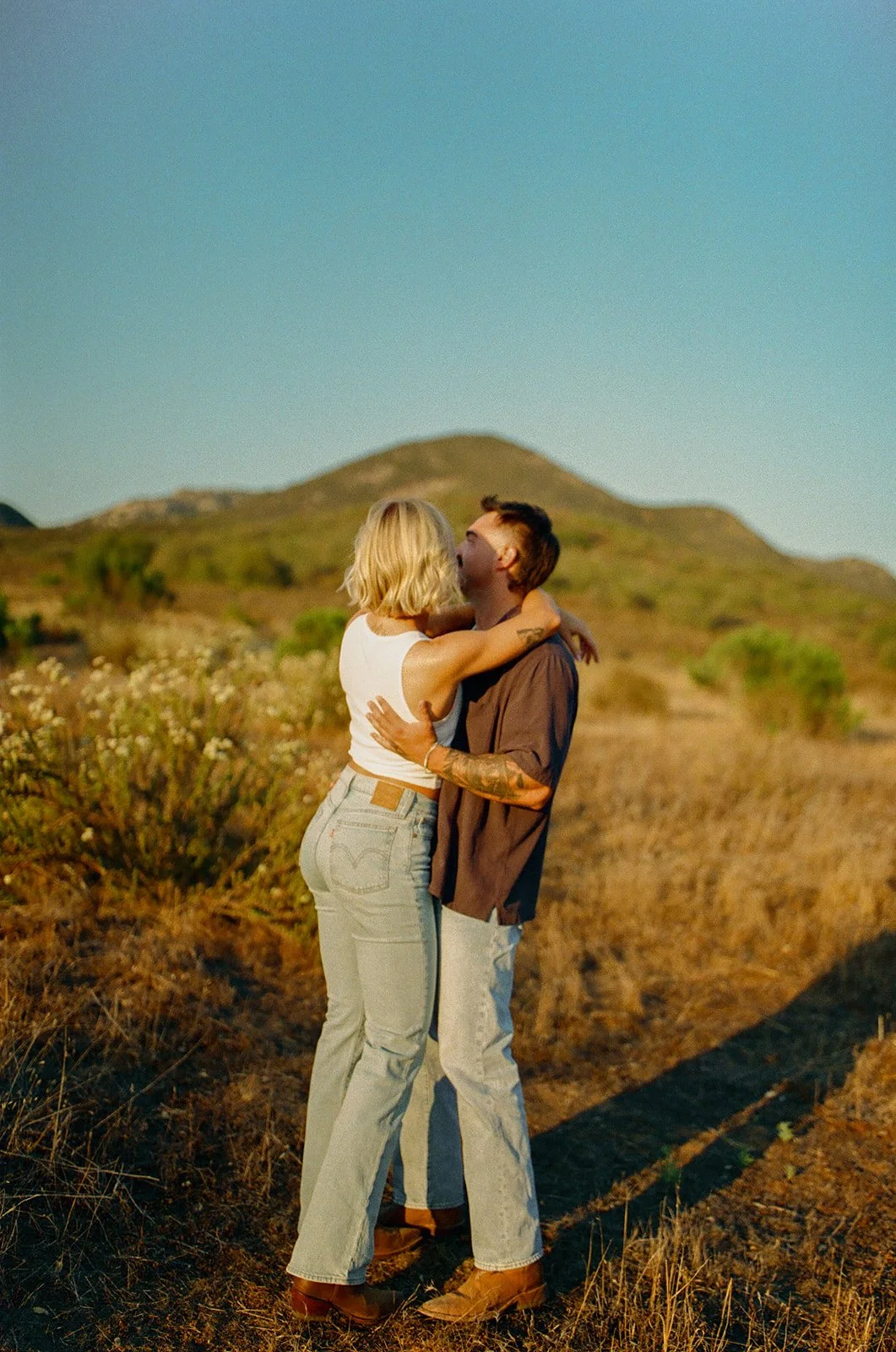 Golden hour western engagement photos of the couple hugging in a mountain field, denim outfits and boots adding to the laid-back vibe.