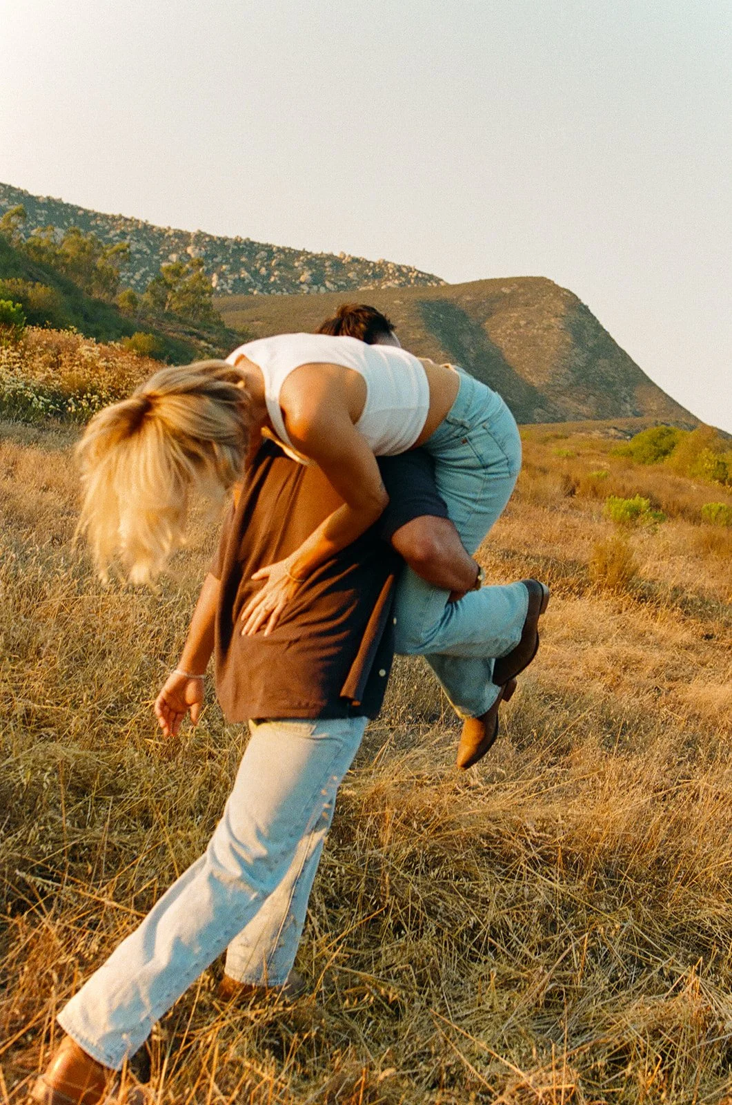 Playful western engagement photos of the guy lifting his fiance up over his shoulder as she laughs in a sunlit field surrounded by rolling hills.
