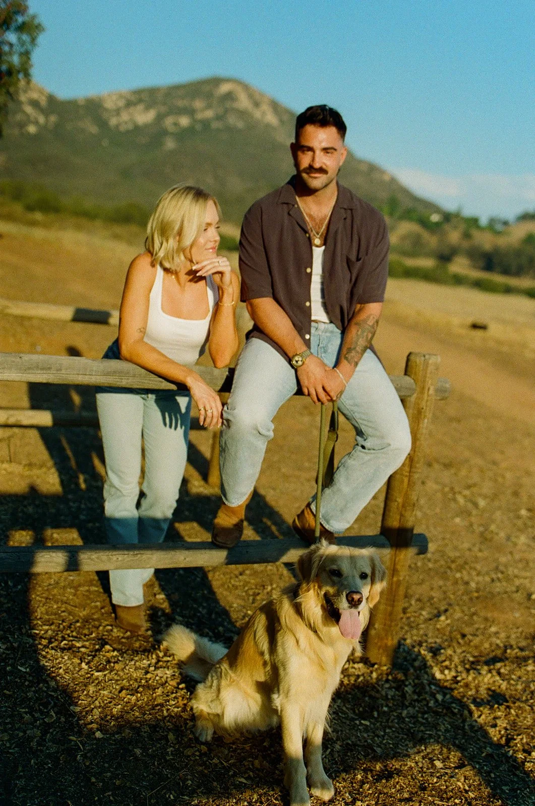Couple sitting on a wooden fence together with their dog below them, mountains stretching across the background.