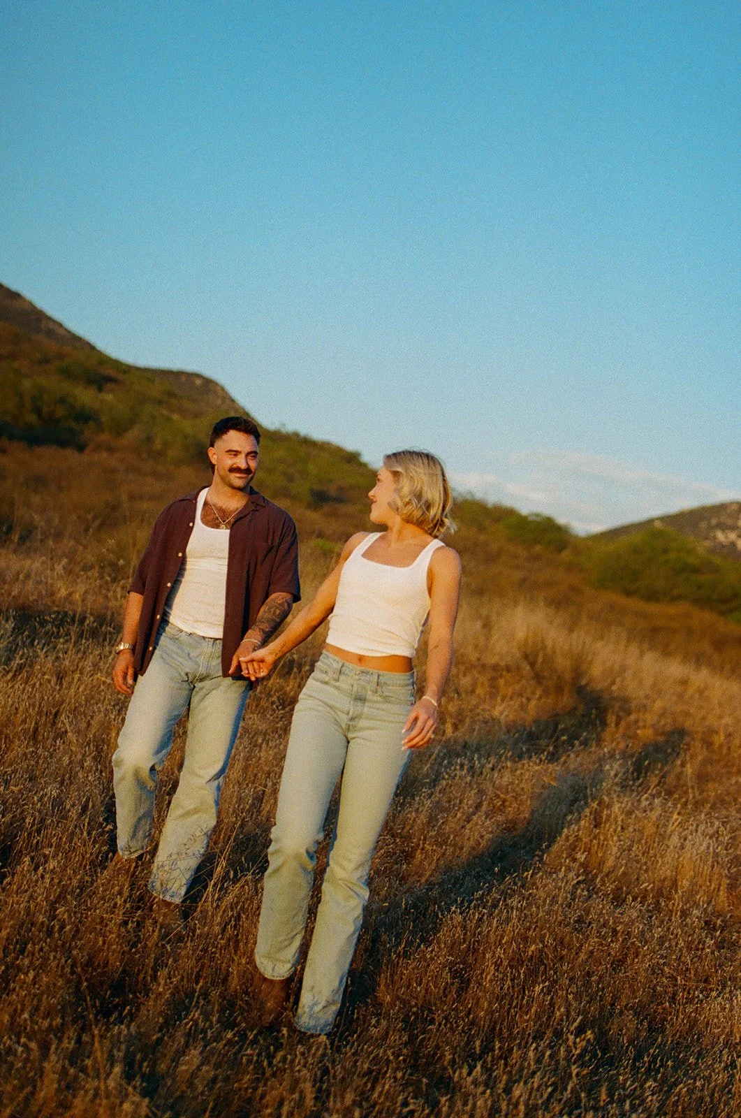 Couple walking hand in hand through a golden field at sunset, wearing denim and boots in a relaxed mountain setting.