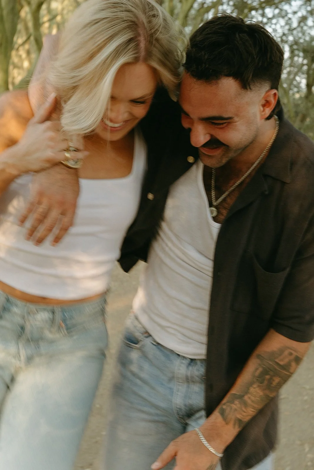 Playful candid moment of the couple bending down to greet their golden retriever on a shaded dirt trail.