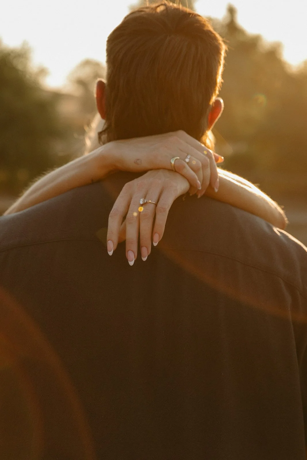 Detail shot of the bride’s arms wrapped around her partner’s shoulders, engagement ring visible in soft sunset light.