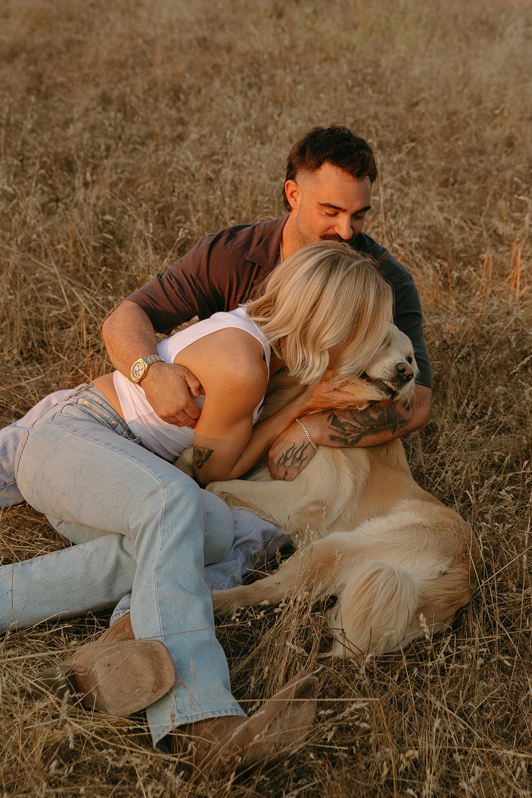 Candid portrait of the couple lying in the grass hugging their golden retriever during golden hour.