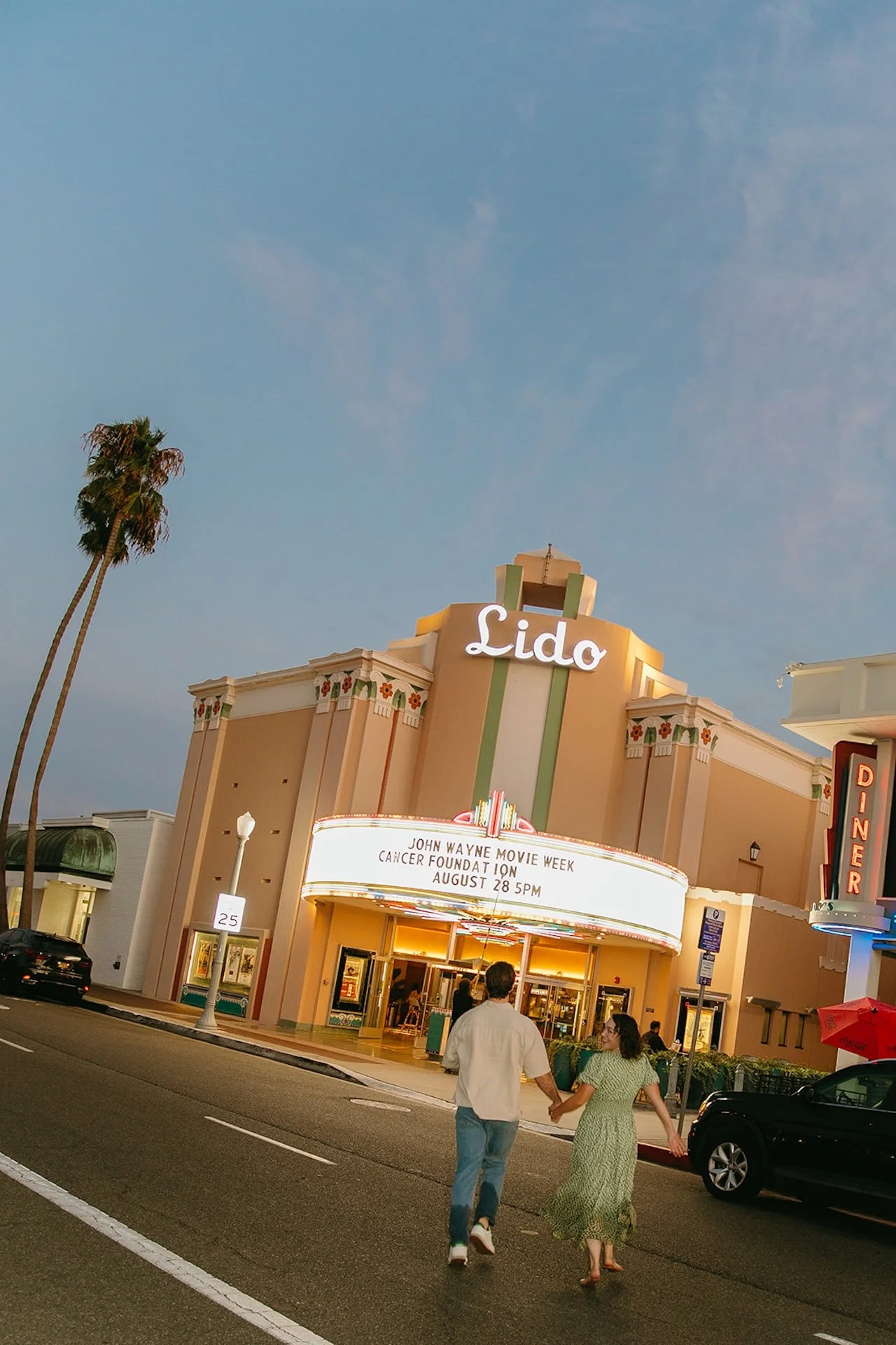 Wide evening photo of a couple holding hands while crossing the street toward Lido Theater, glowing marquee lights and palm trees framing the scene.