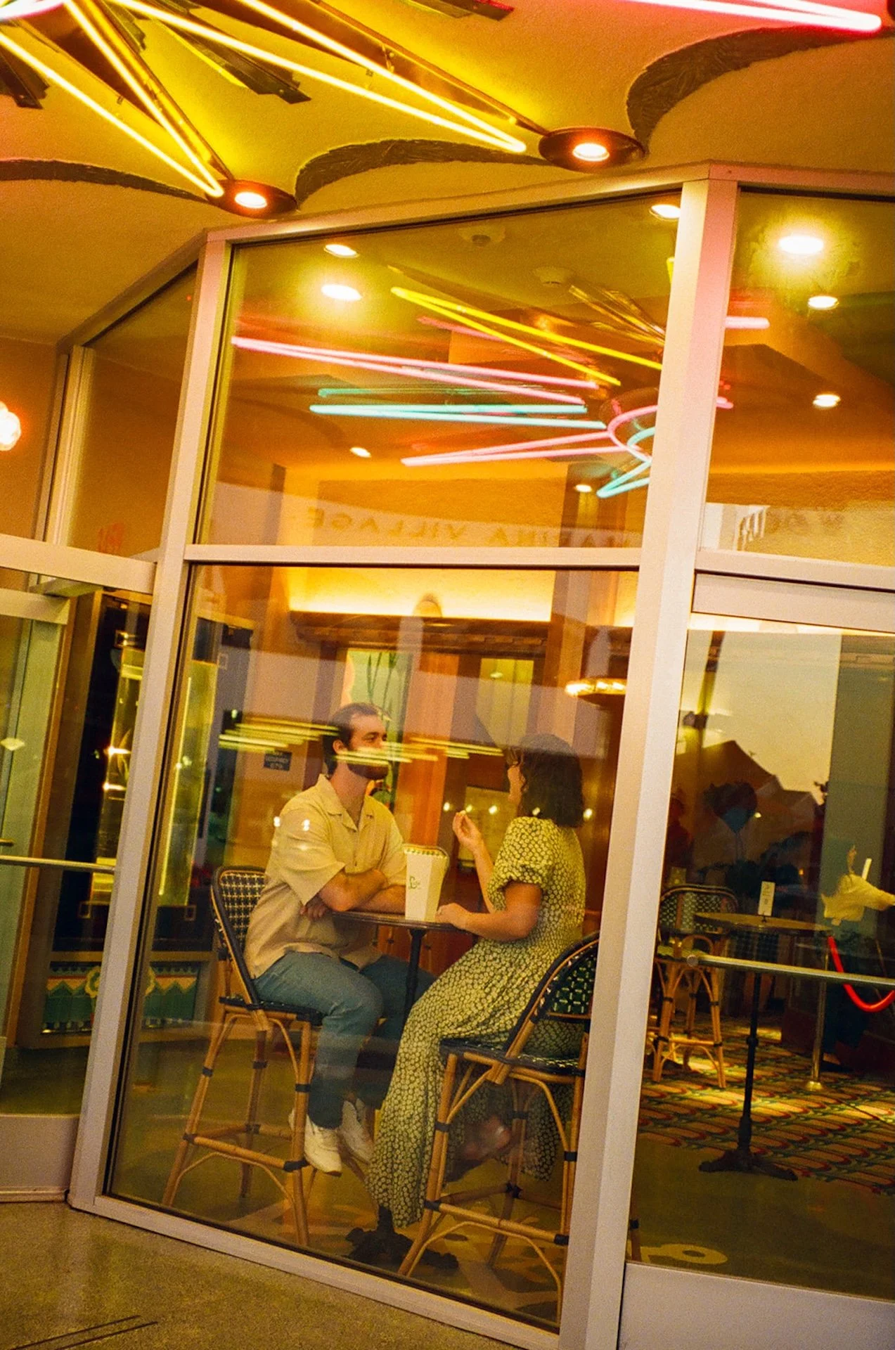Couple sitting inside a retro diner window at Lido Village, neon lights reflecting on the glass as they talk across a small table.