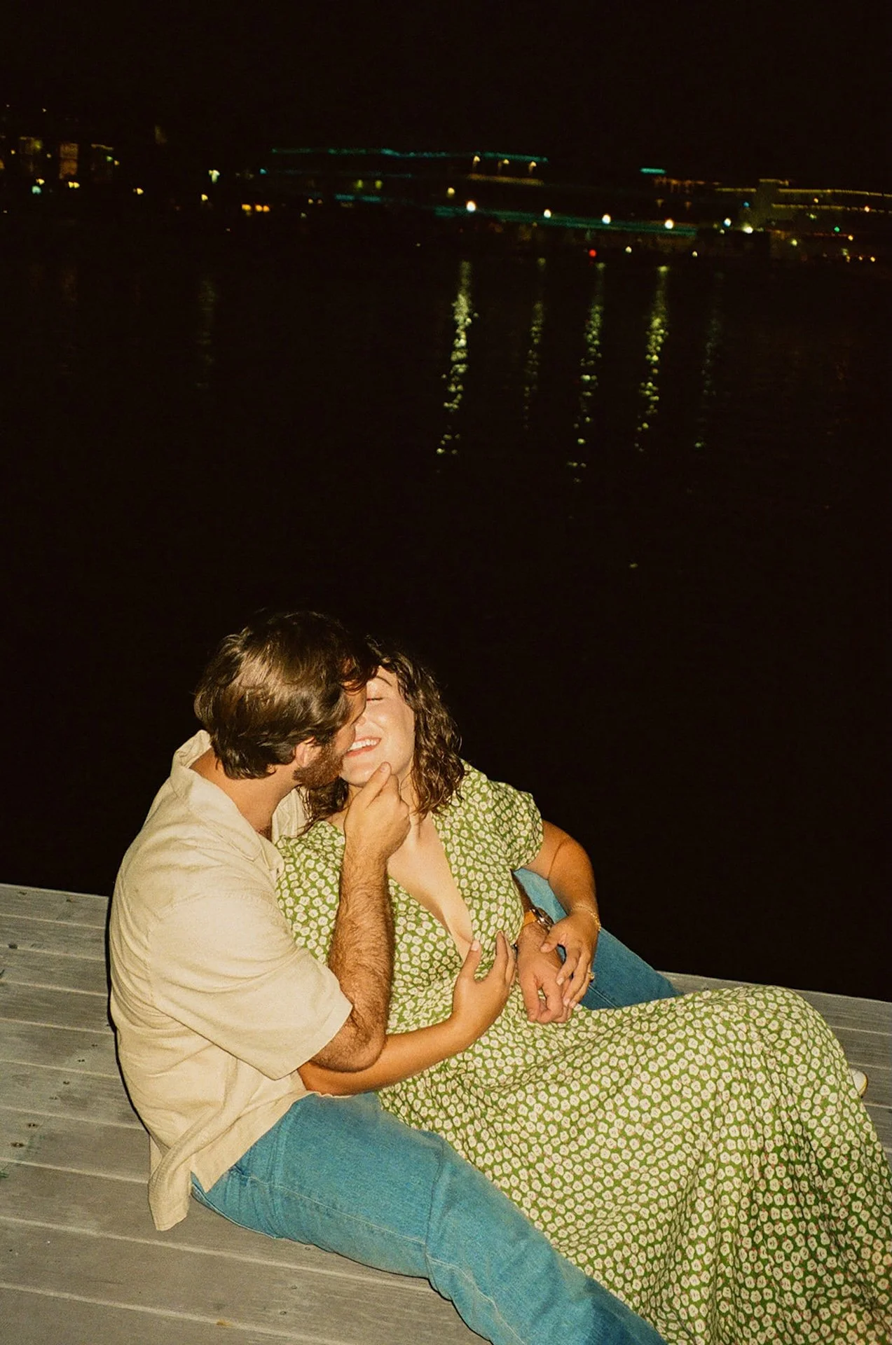Nighttime candid of a couple sitting on a dock by the water, laughing and cuddling under city lights reflecting across the harbor.