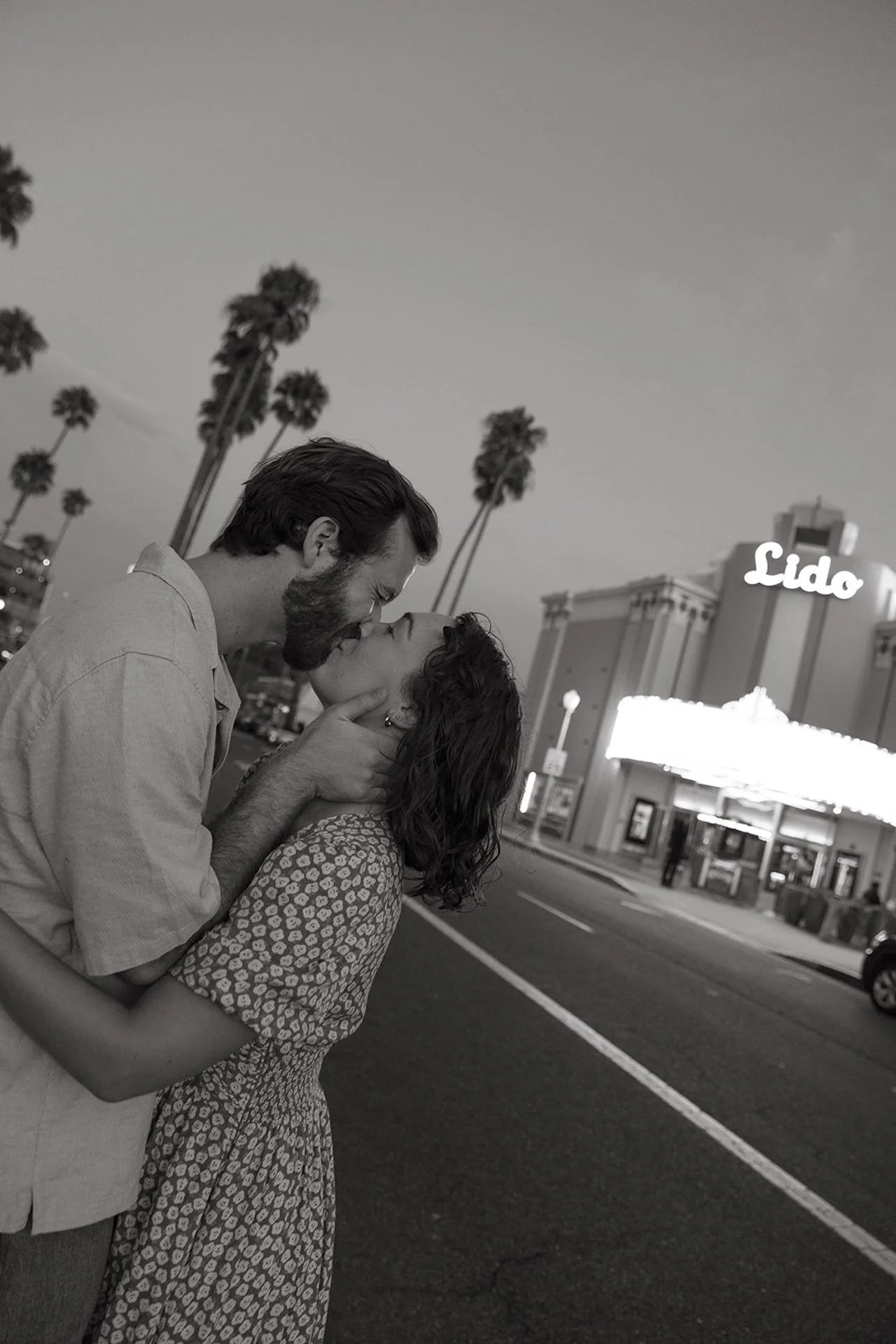 Black and white image of a couple kissing in the street outside Lido Theater in Newport Beach with palm trees and vintage marquee lights behind them.