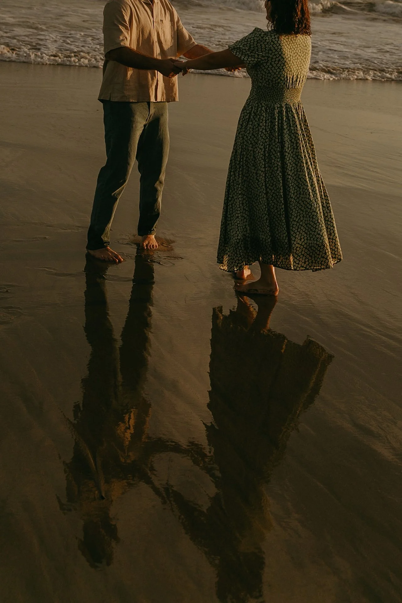 Couple holding hands barefoot in the wet sand at the shoreline, their reflections glowing in the warm evening light.