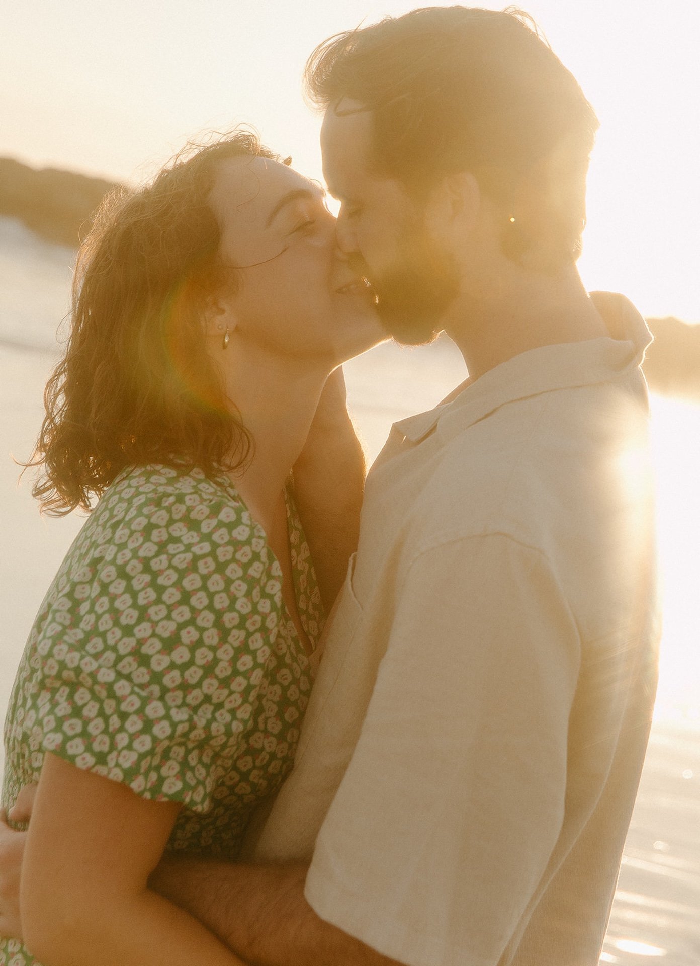Close-up of a couple kissing at sunset during their newport beach engagement photos, golden light flaring around them as waves roll in behind.