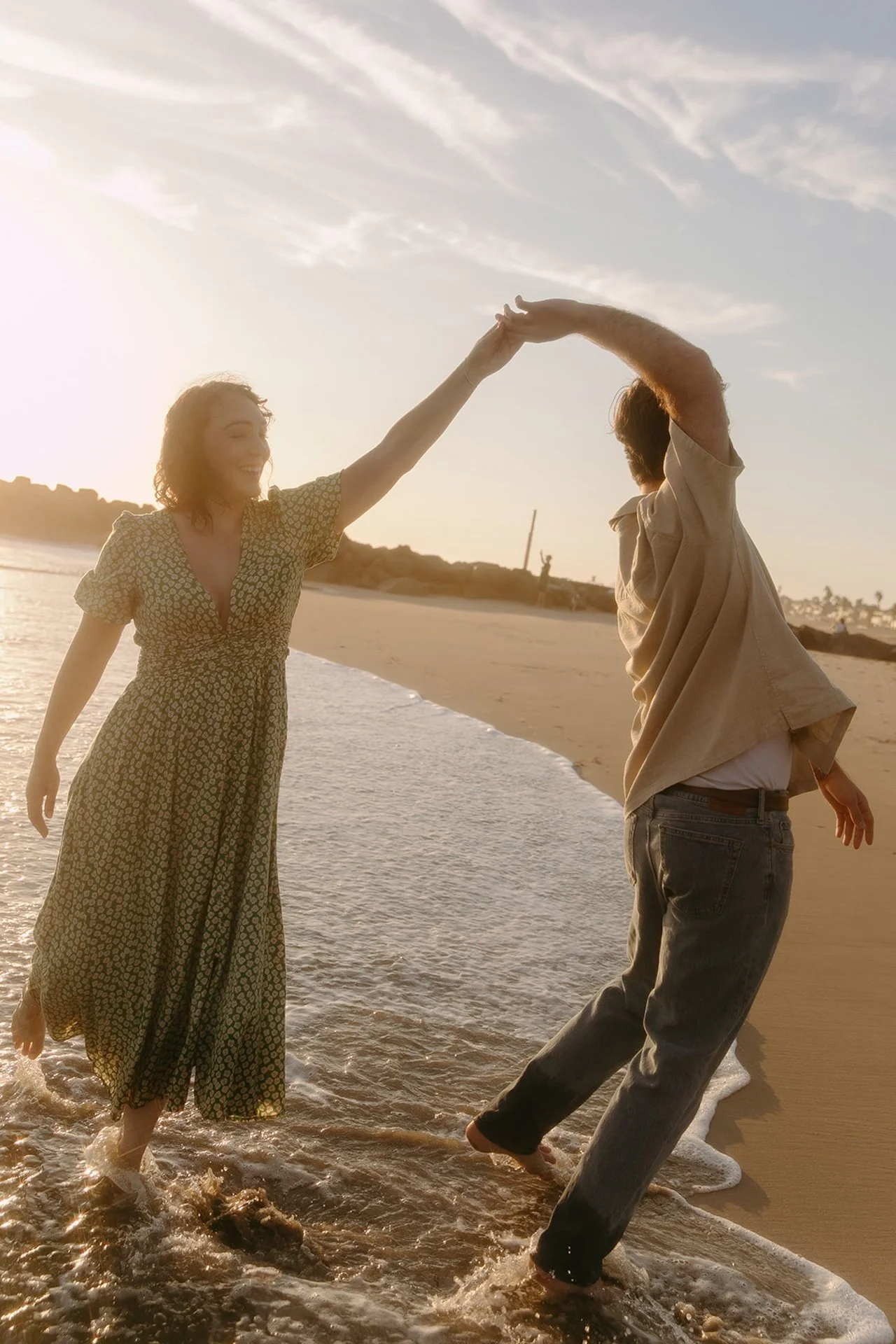 Couple holding hands and dancing barefoot in the wet sand at the shoreline, their reflections glowing in the warm evening light.
