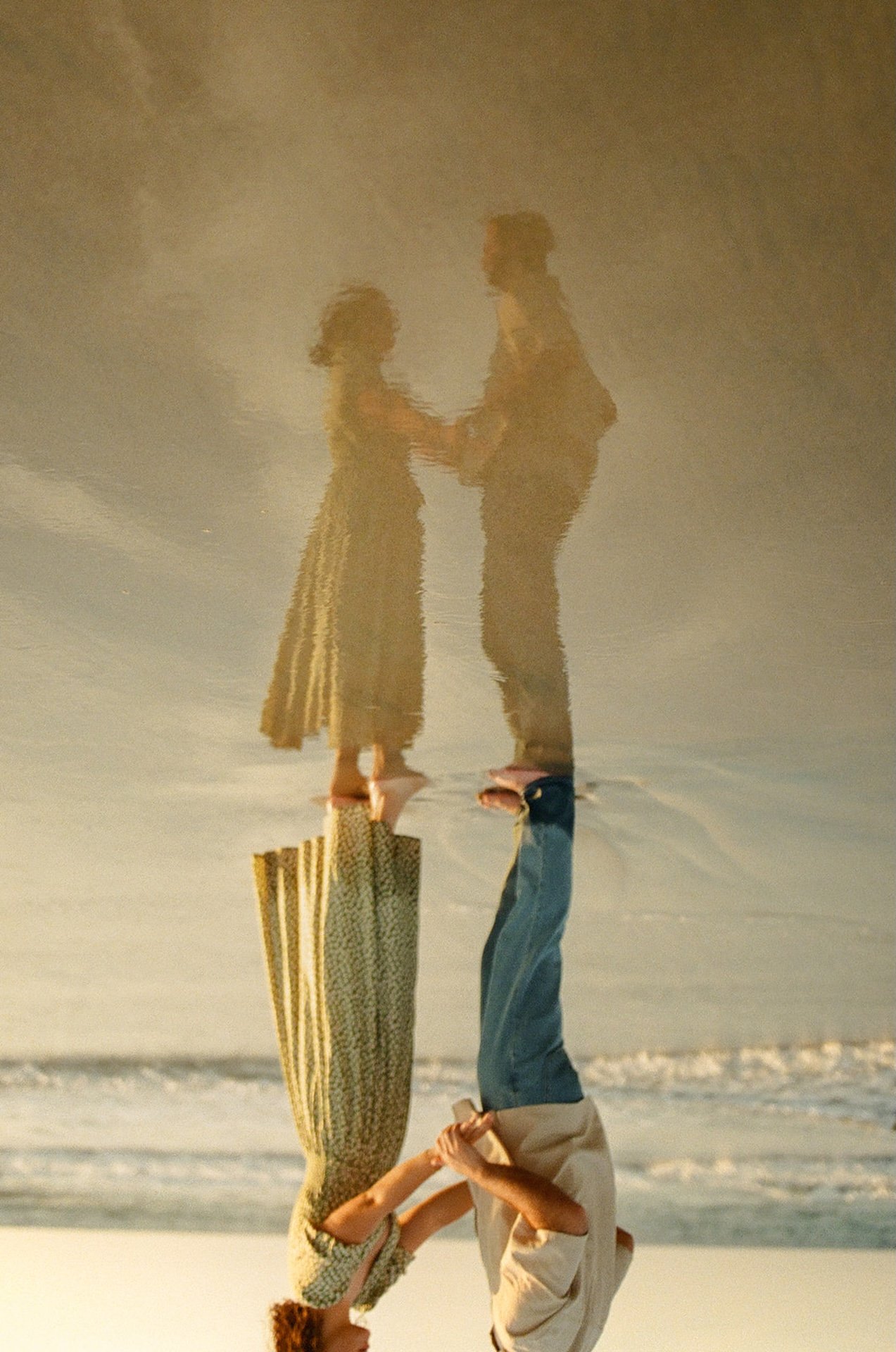An artistic engagement shoot photo of the couple holding hands on the beach with their reflection in the sand.