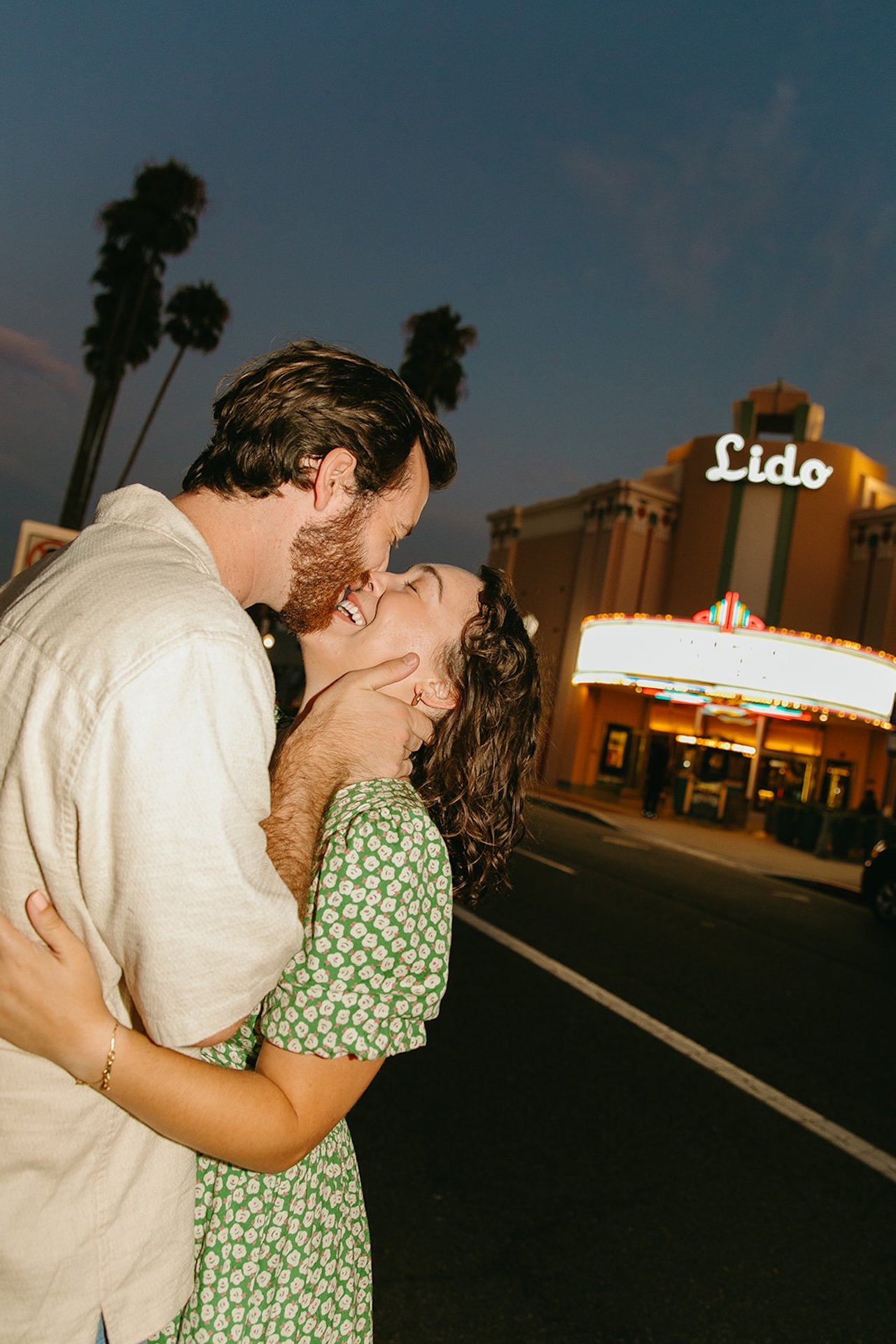 Romantic photo of a couple kissing in the street outside Lido Theater in Newport Beach with palm trees and vintage marquee lights behind them.