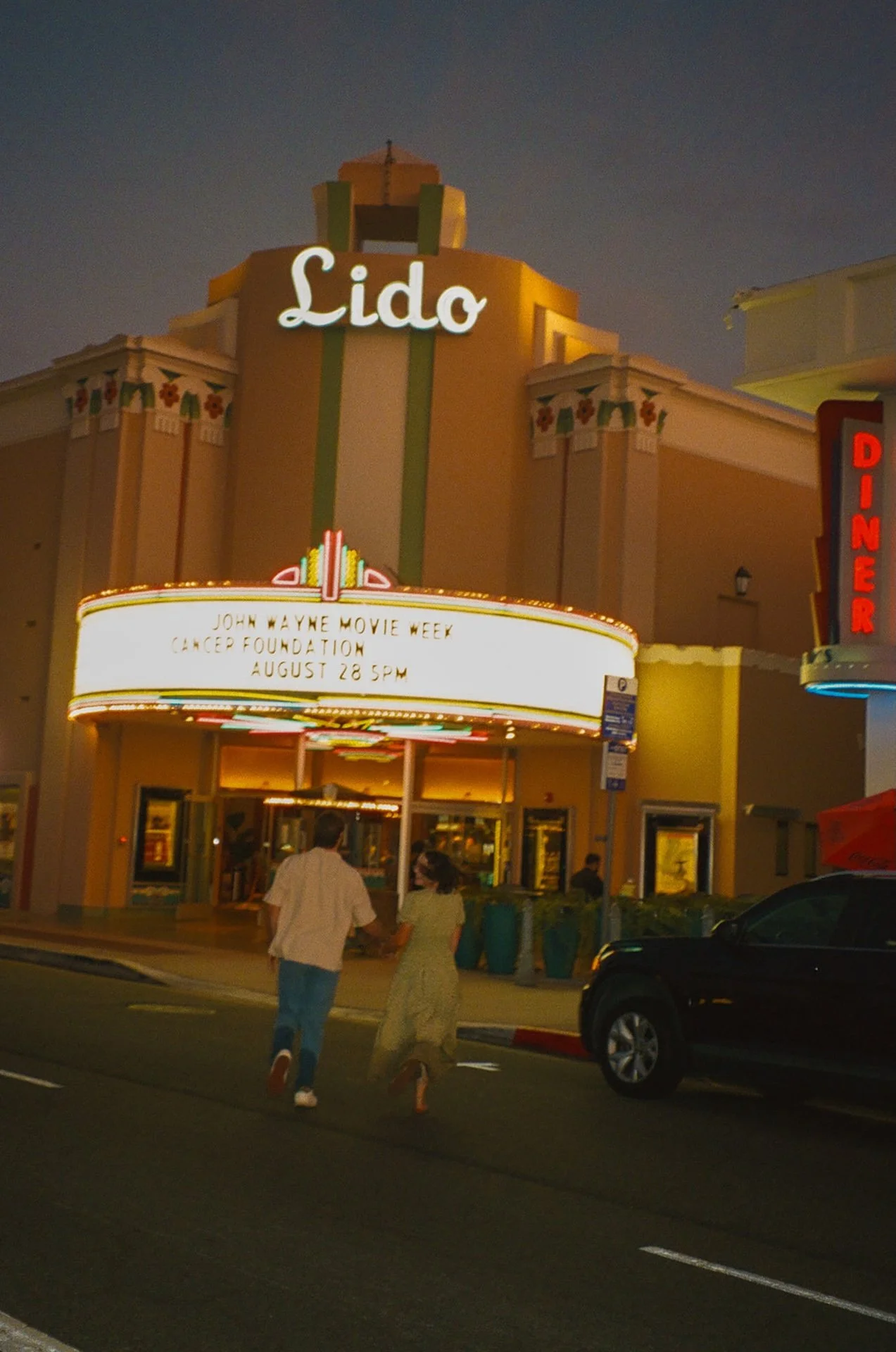 Wide evening photo of a couple holding hands while crossing the street toward Lido Theater, glowing marquee lights and palm trees framing the scene.