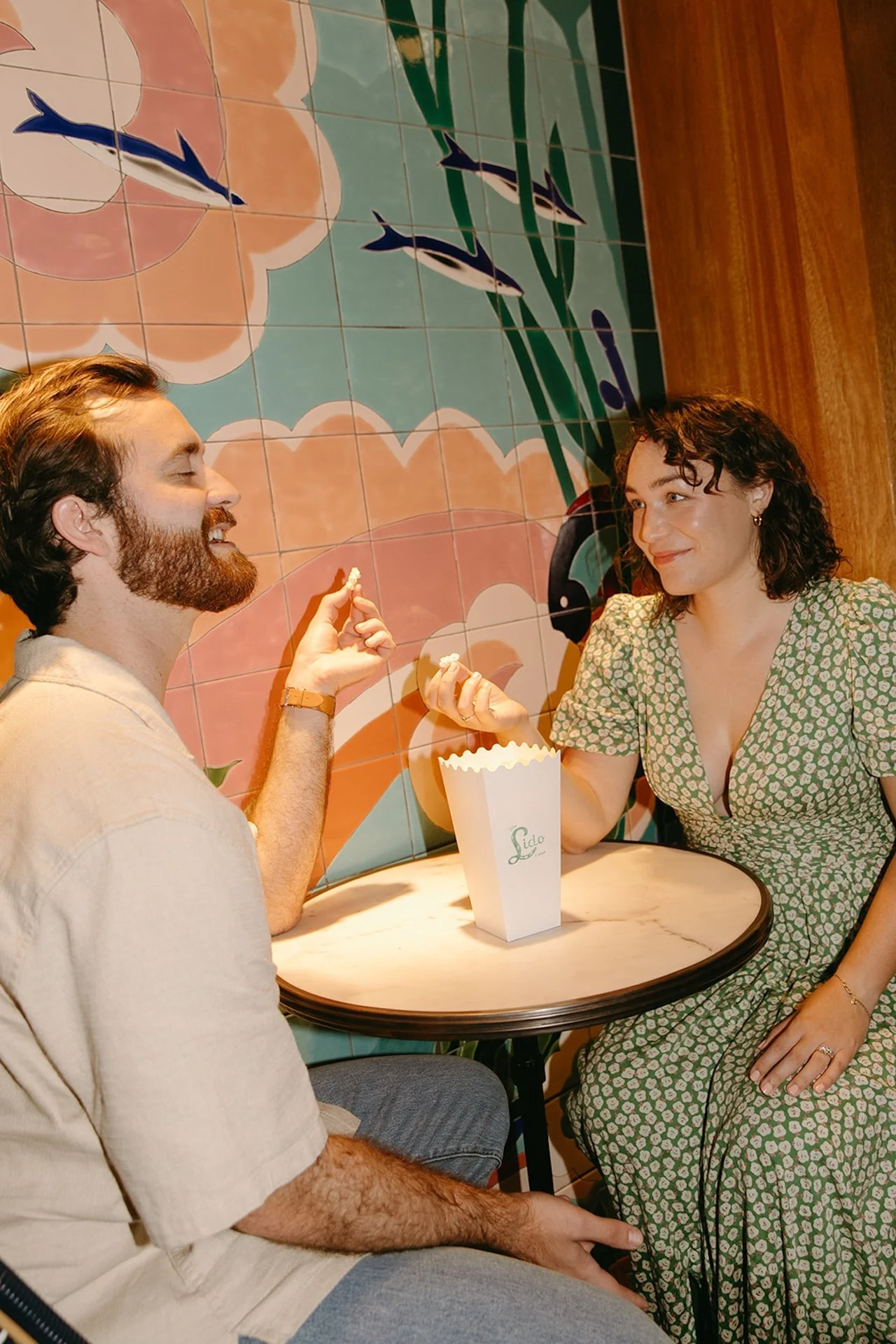 A couple sitting at a small table sharing popcorn during their engagement shoot.