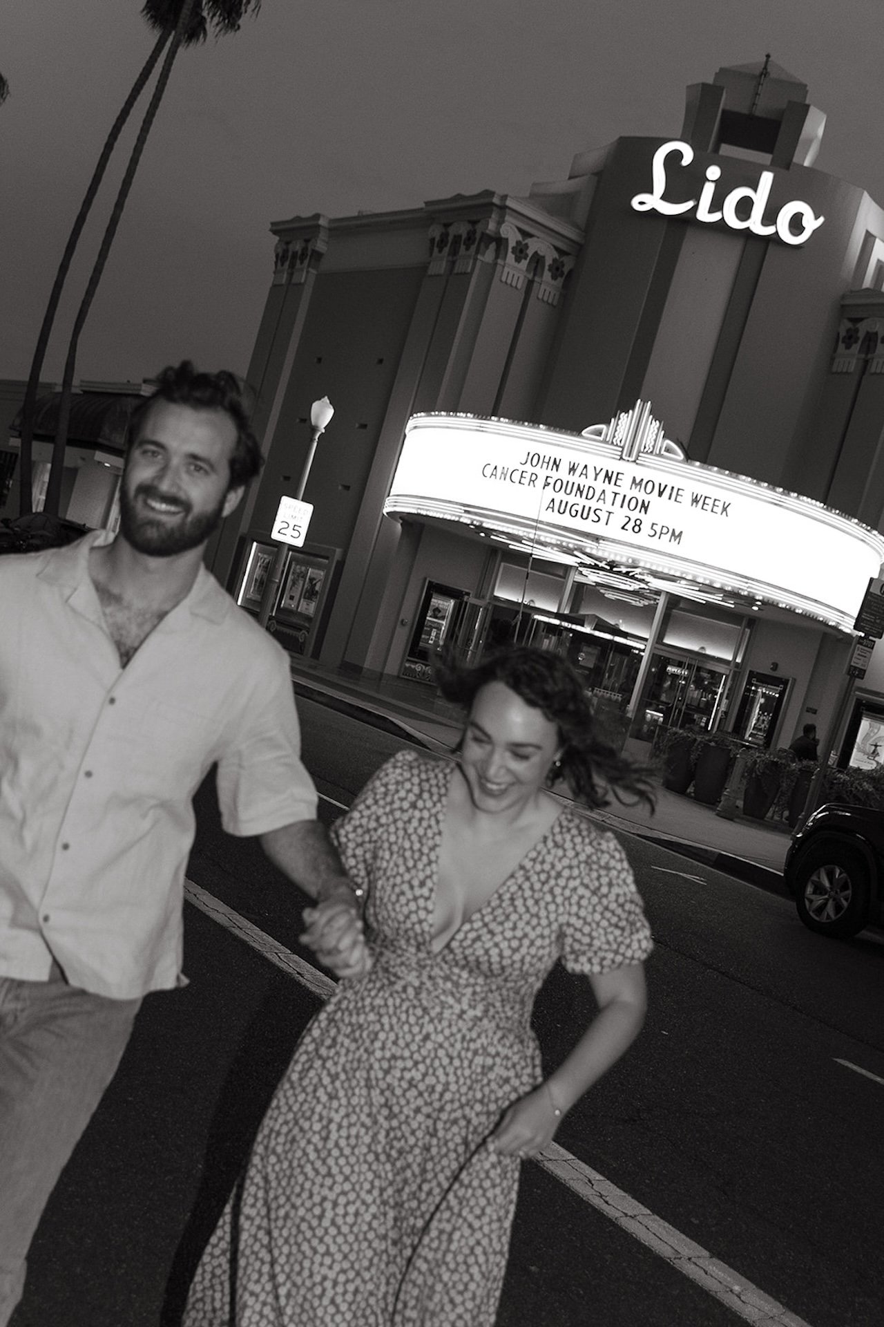 A candid photo of a couple walking towards the camera with the Lido theater behind them during their Newport Beach engagement photos
