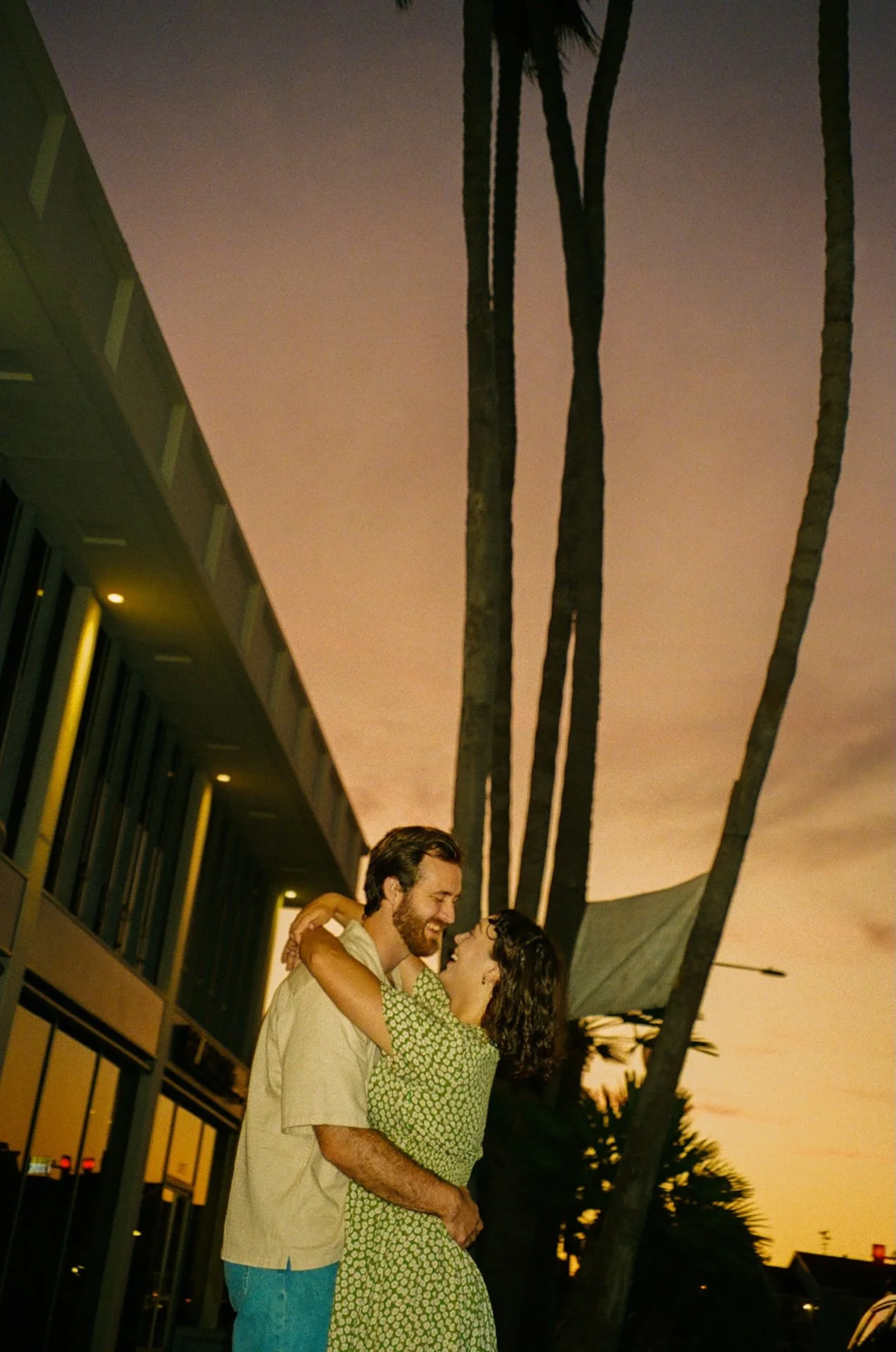 A candid photo of a couple hugging and sharing a kiss with the sunset and palm trees behind them.