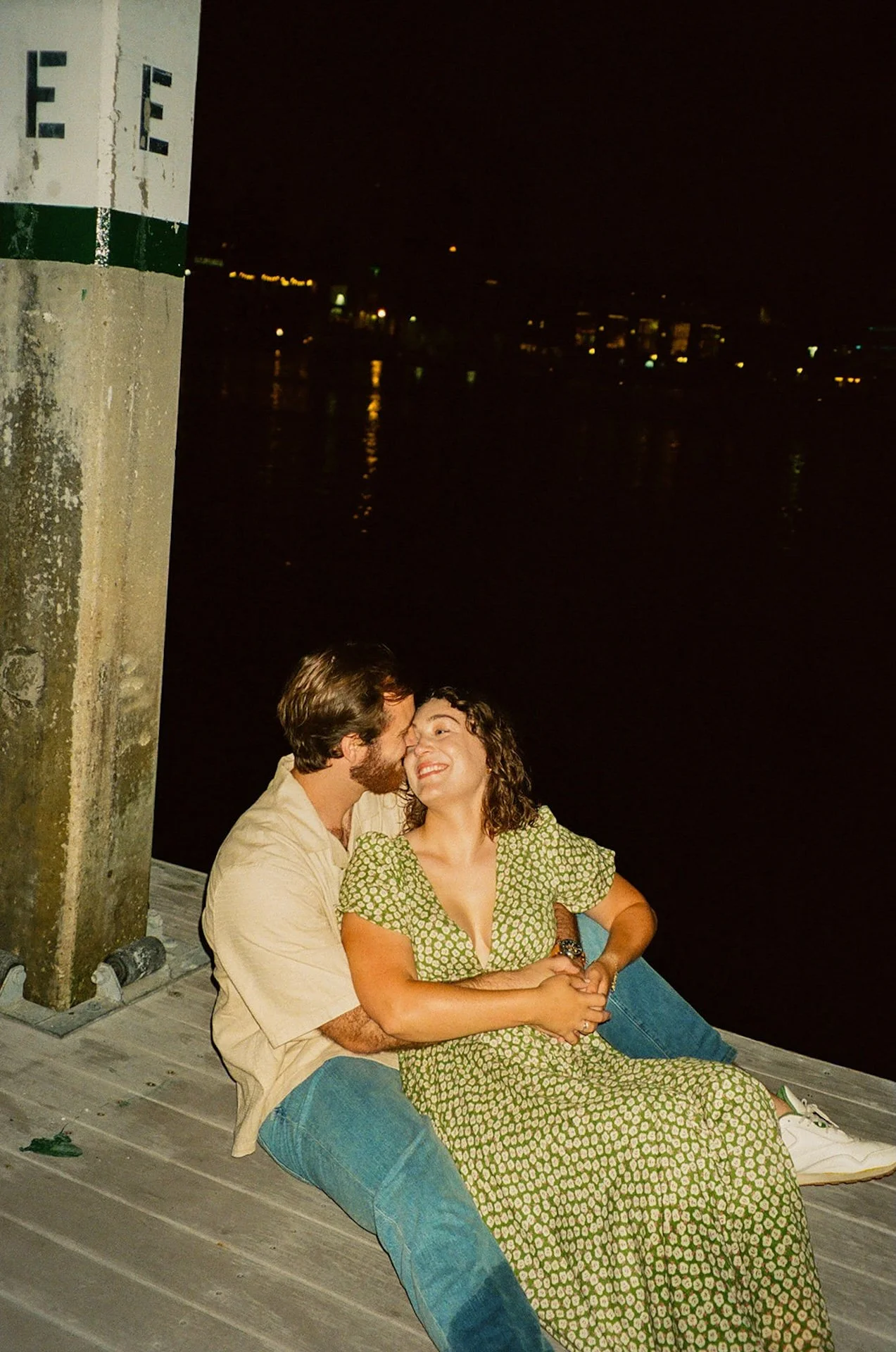 Nighttime candid flash photo of a couple leaning into each other on a dock by the water, with the city lights reflecting across the harbor.