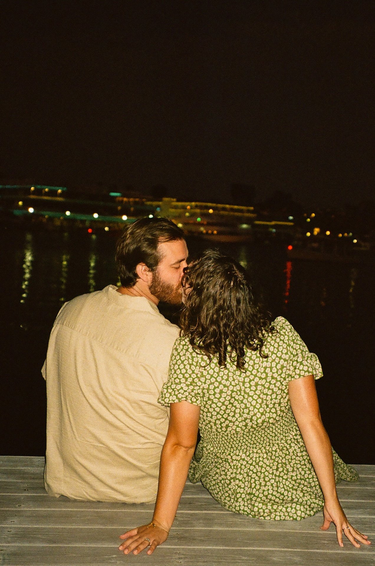 Nighttime candid flash photo of a couple sitting on a dock by the water, kissing and cuddling under city lights reflecting across the harbor.