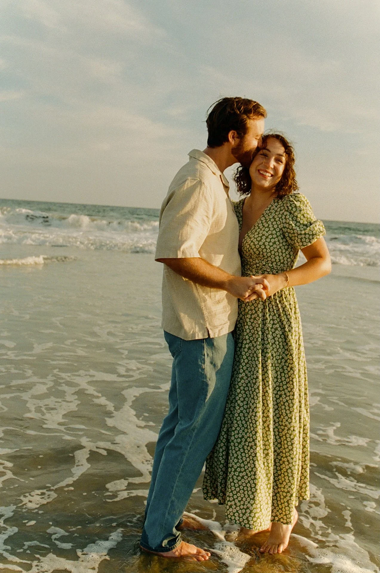 An engaged couple holding hands while the waves crash against their feet. 