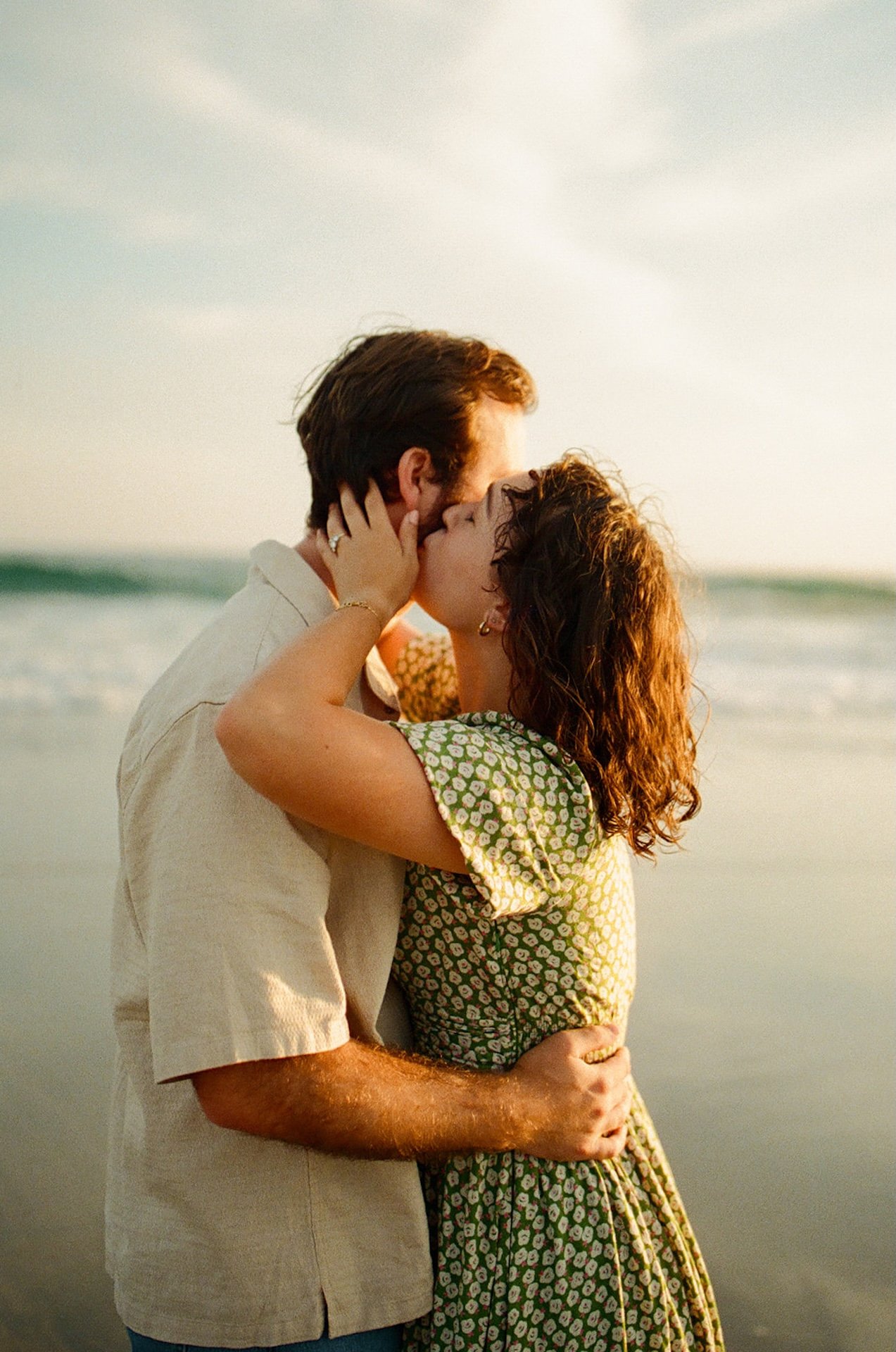 A photo of the girl kissing her fiance during their engagement shoot with the sunset and the beach in the background.