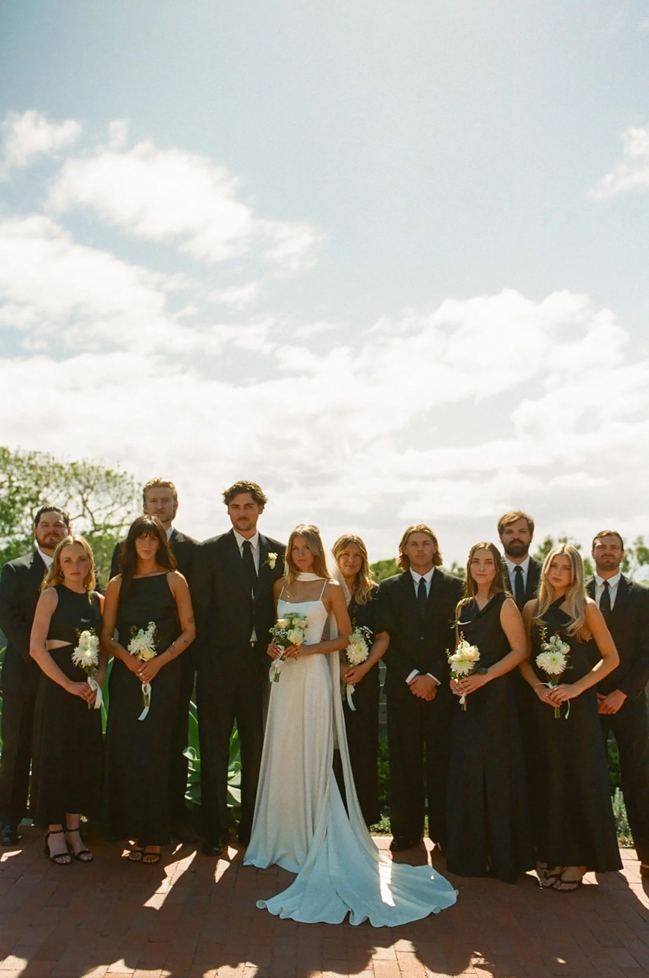 Wedding party standing together outdoors in black attire with white florals during a sunny coastal ceremony portrait.