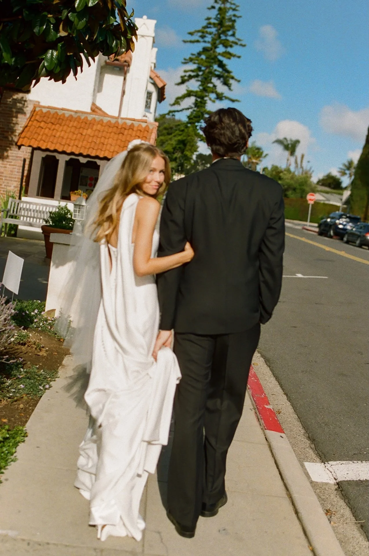 Bride looking back and smiling while walking arm in arm with the groom outside a Spanish-style church in Laguna Beach.