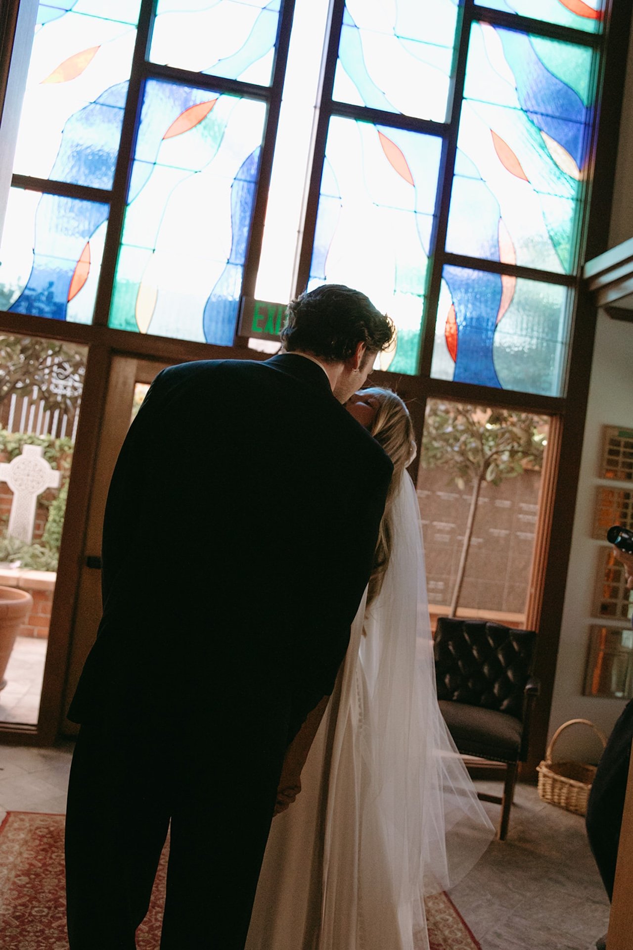 Bride and groom sharing a quiet kiss beneath colorful stained glass windows inside the church entryway.