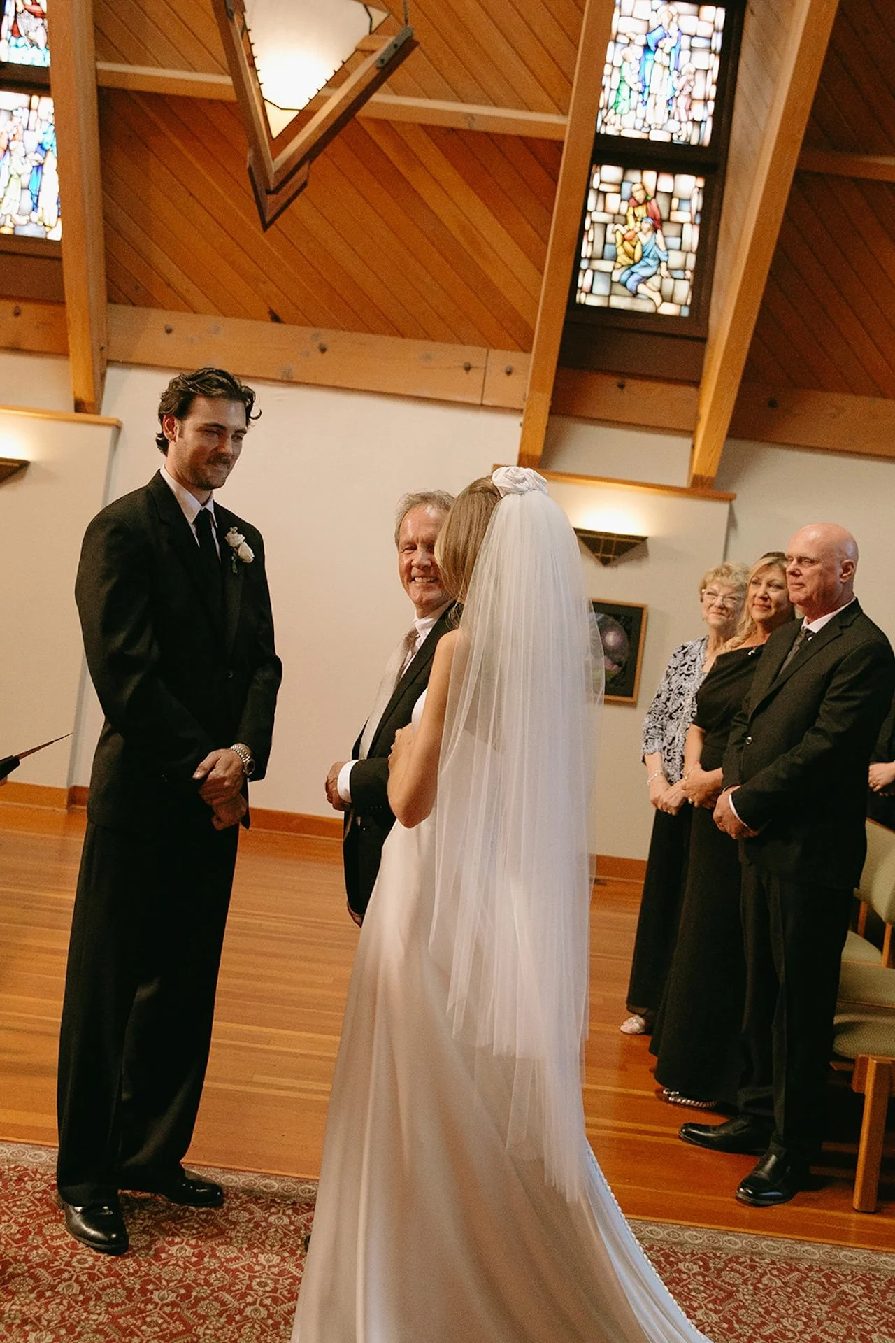 Bride standing with her father during the ceremony while family members watch inside a warm wood chapel with stained glass windows.