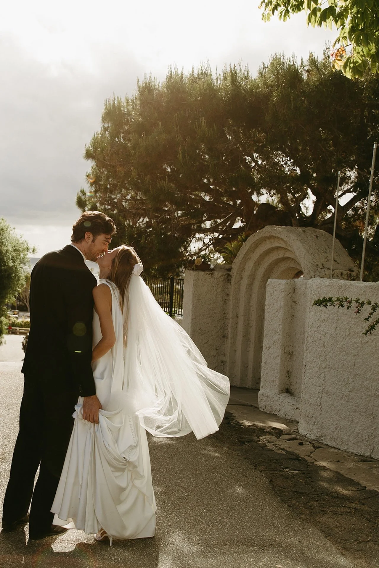 Bride and groom sharing a romantic kiss on a quiet Laguna Beach street with her veil flowing in the breeze.