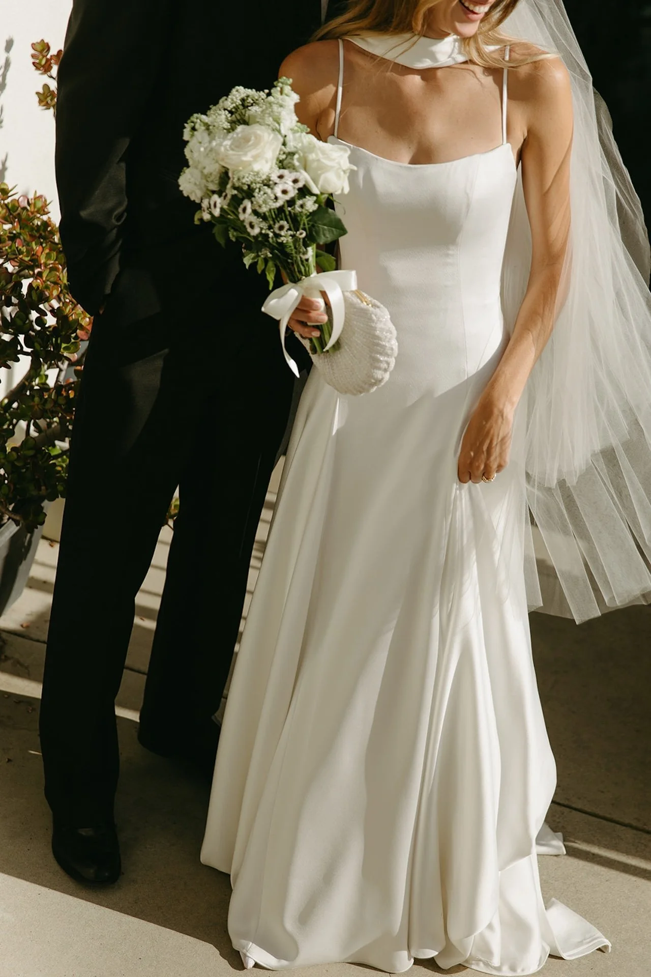 Close-up of the bride’s minimalist satin wedding dress, bouquet of white flowers, and delicate veil in soft golden sunlight.