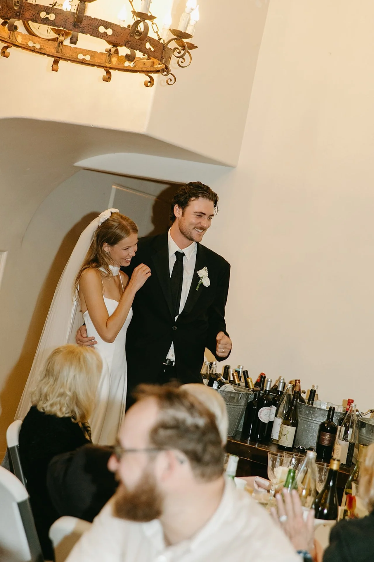 Bride and groom entering their wedding reception smiling and holding each other close beside a table filled with wine bottles and glasses.