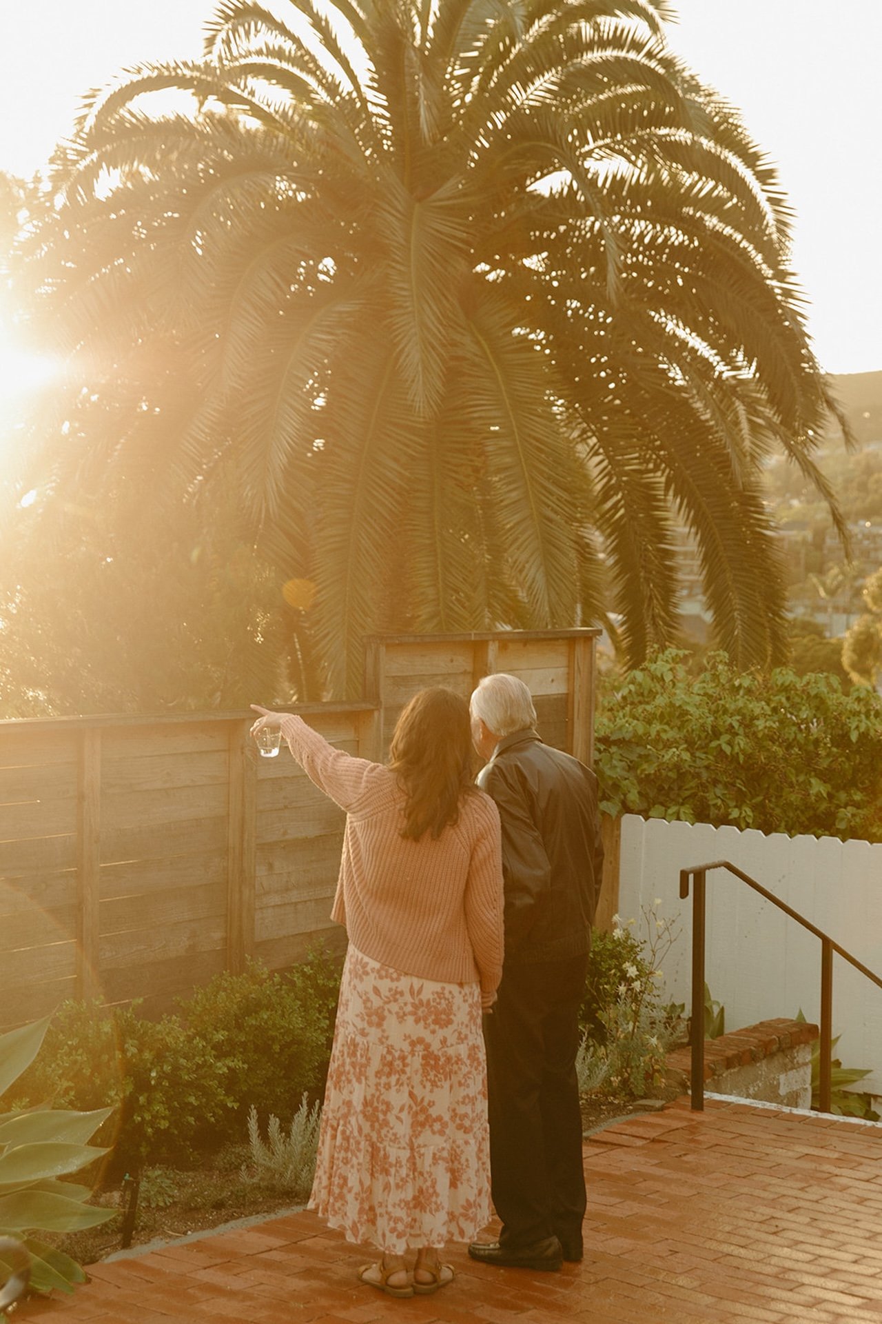 Two guests standing together on a brick patio at sunset beneath a large palm tree, pointing toward the ocean during a relaxed Laguna Beach wedding evening.