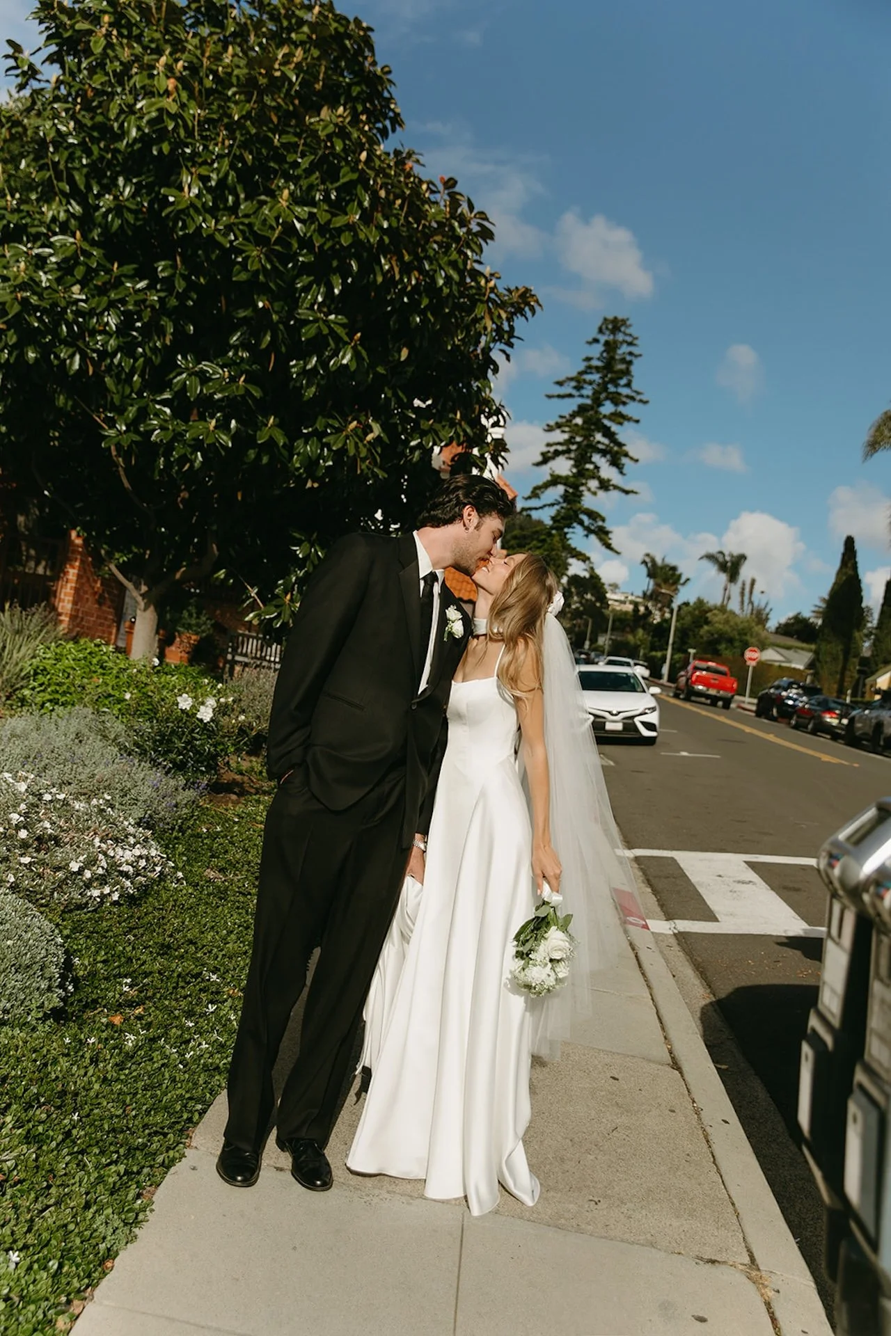 Bride and groom sharing a kiss on a sunny neighborhood sidewalk during romantic Laguna Beach wedding portraits.