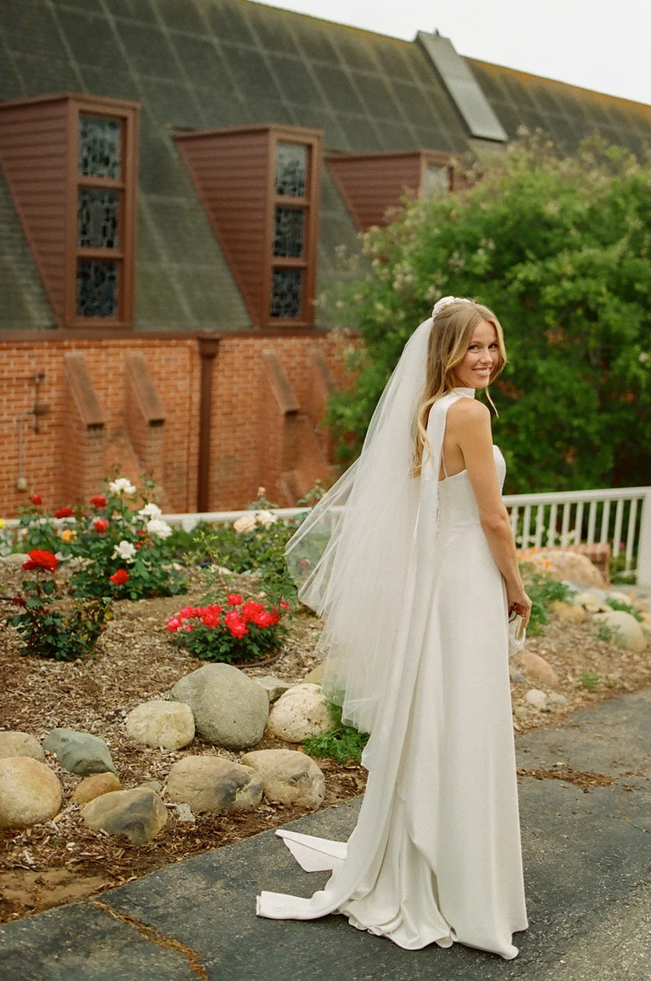 Bride looking back and smiling while standing beside a garden of roses outside the church after her Laguna Beach wedding ceremony.