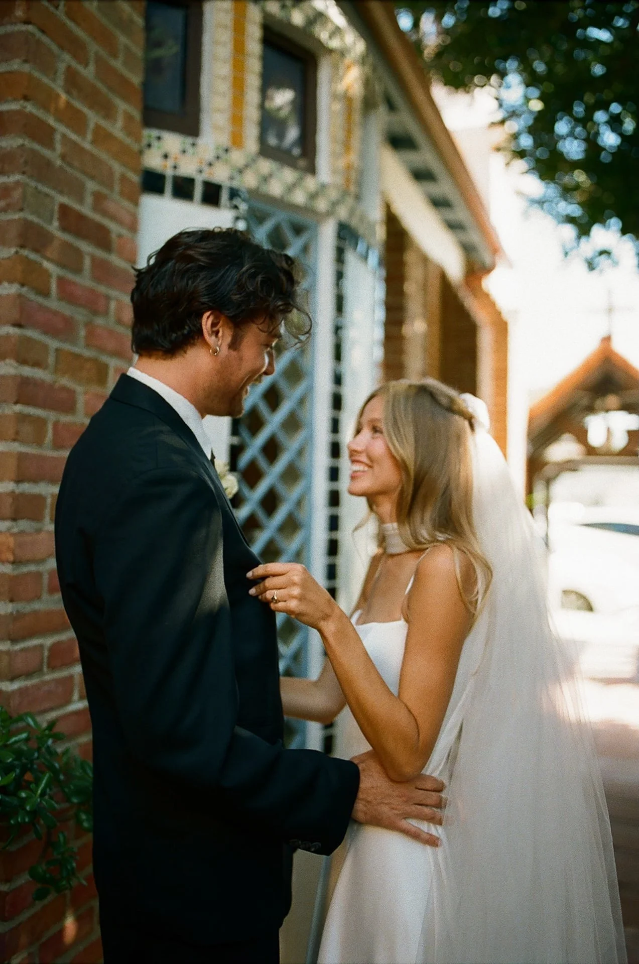 Bride and groom laughing together beside a brick wall and stained glass window during their Laguna Beach wedding portraits.