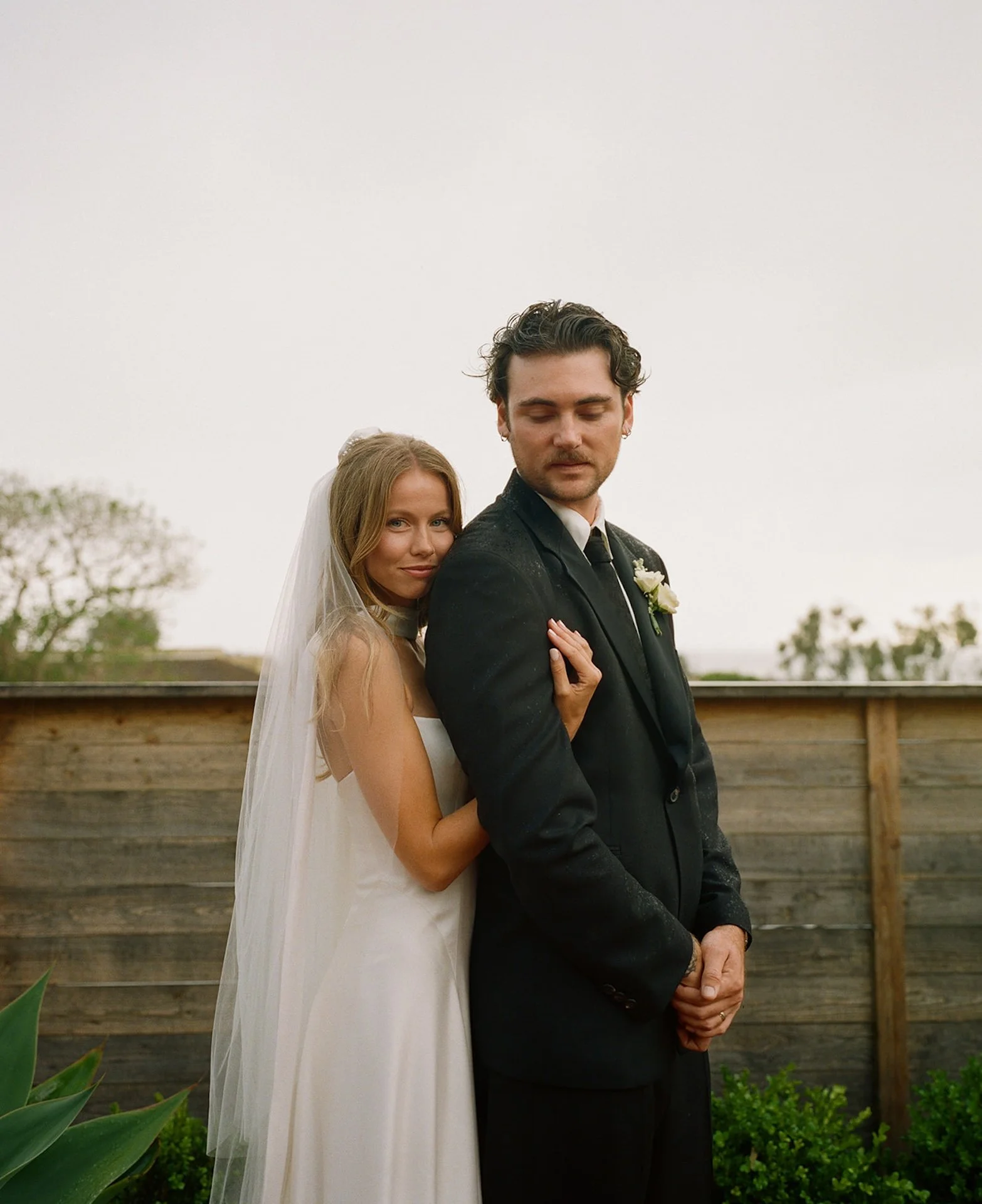 Bride standing behind the groom with her arms around him during romantic Laguna Beach wedding portraits in soft evening light.
