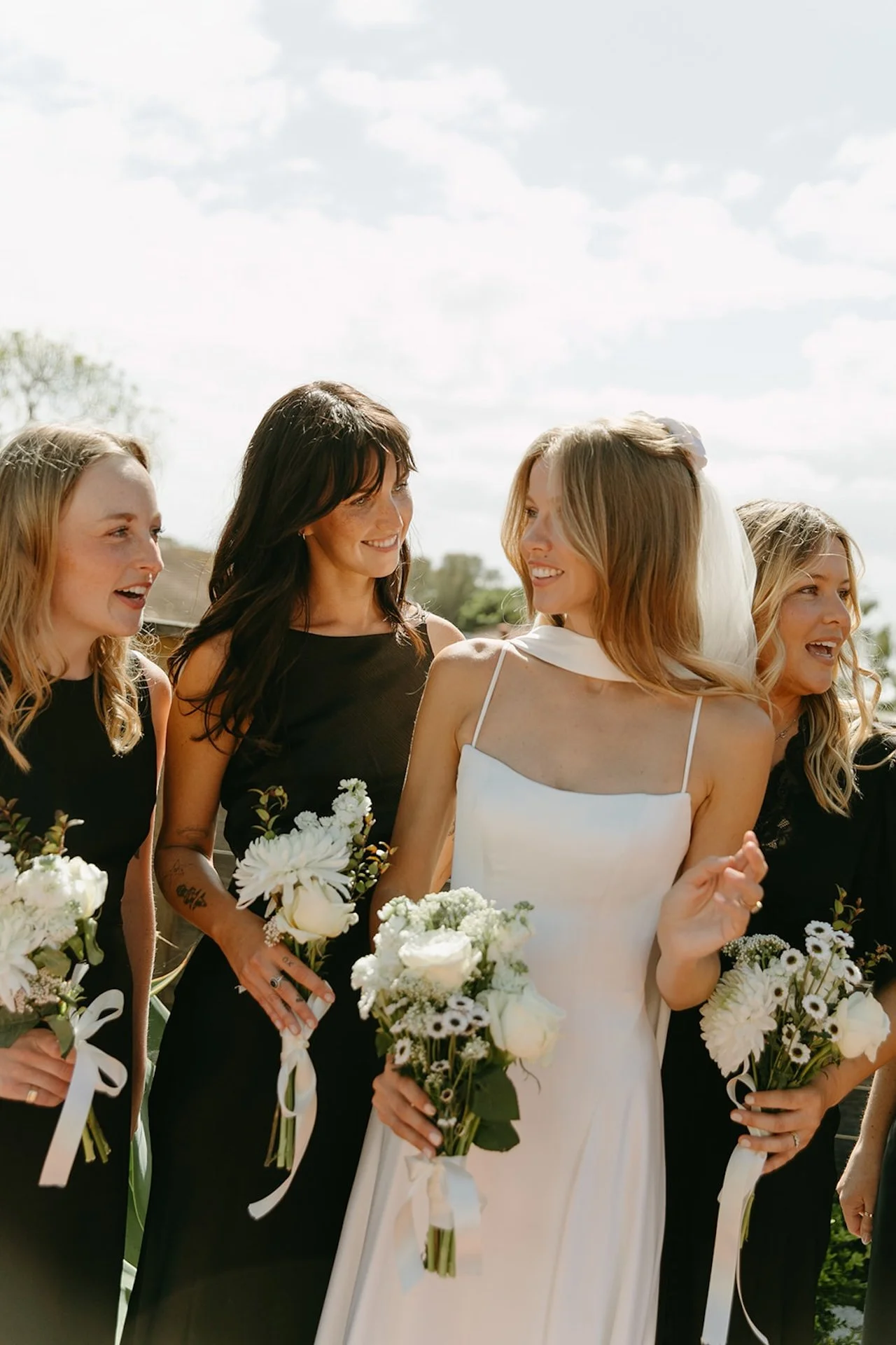 Bride laughing with bridesmaids holding white flower bouquets during candid wedding party portraits.