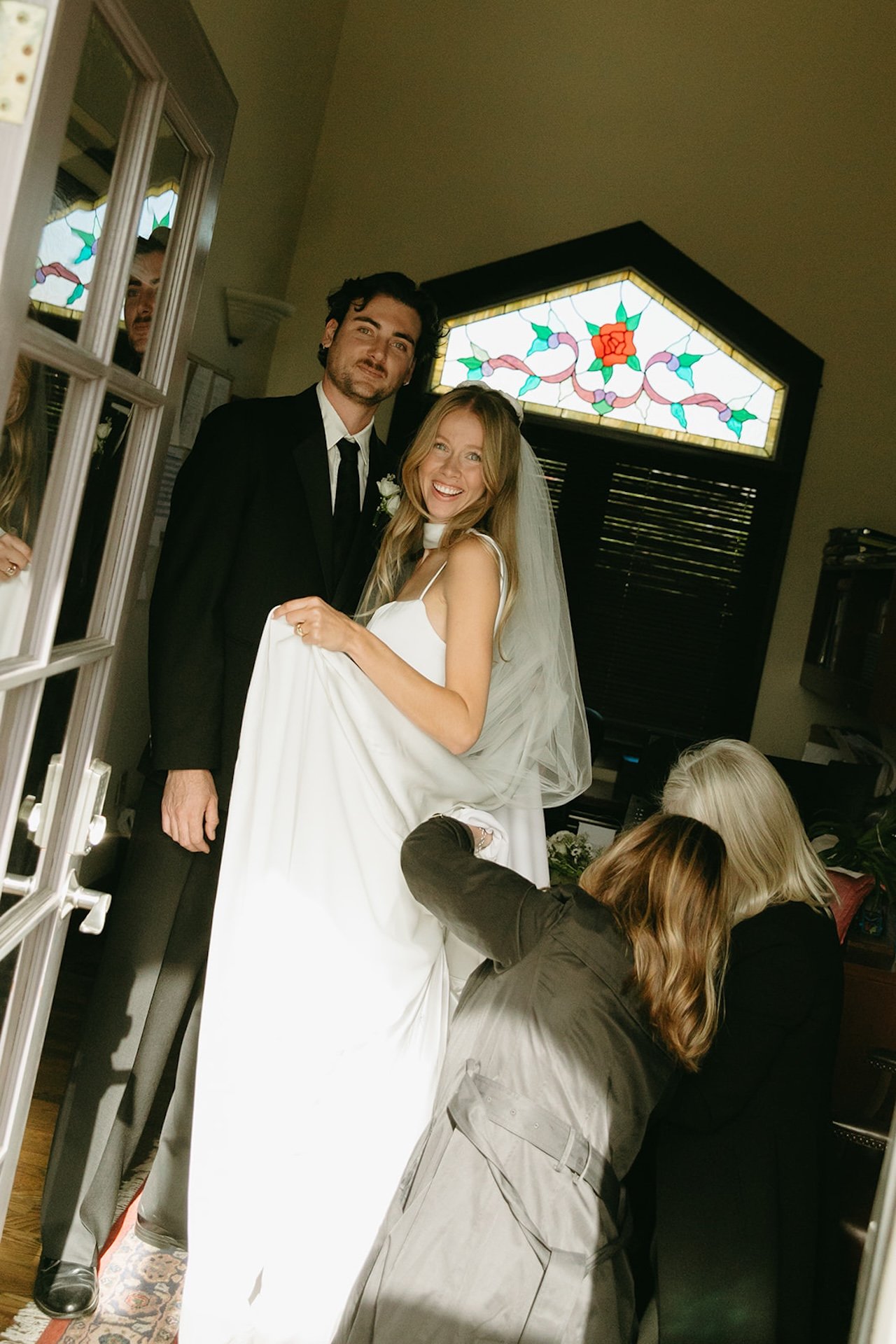 Bride smiling while someone adjusts her dress in a small room beside stained glass windows before the ceremony.