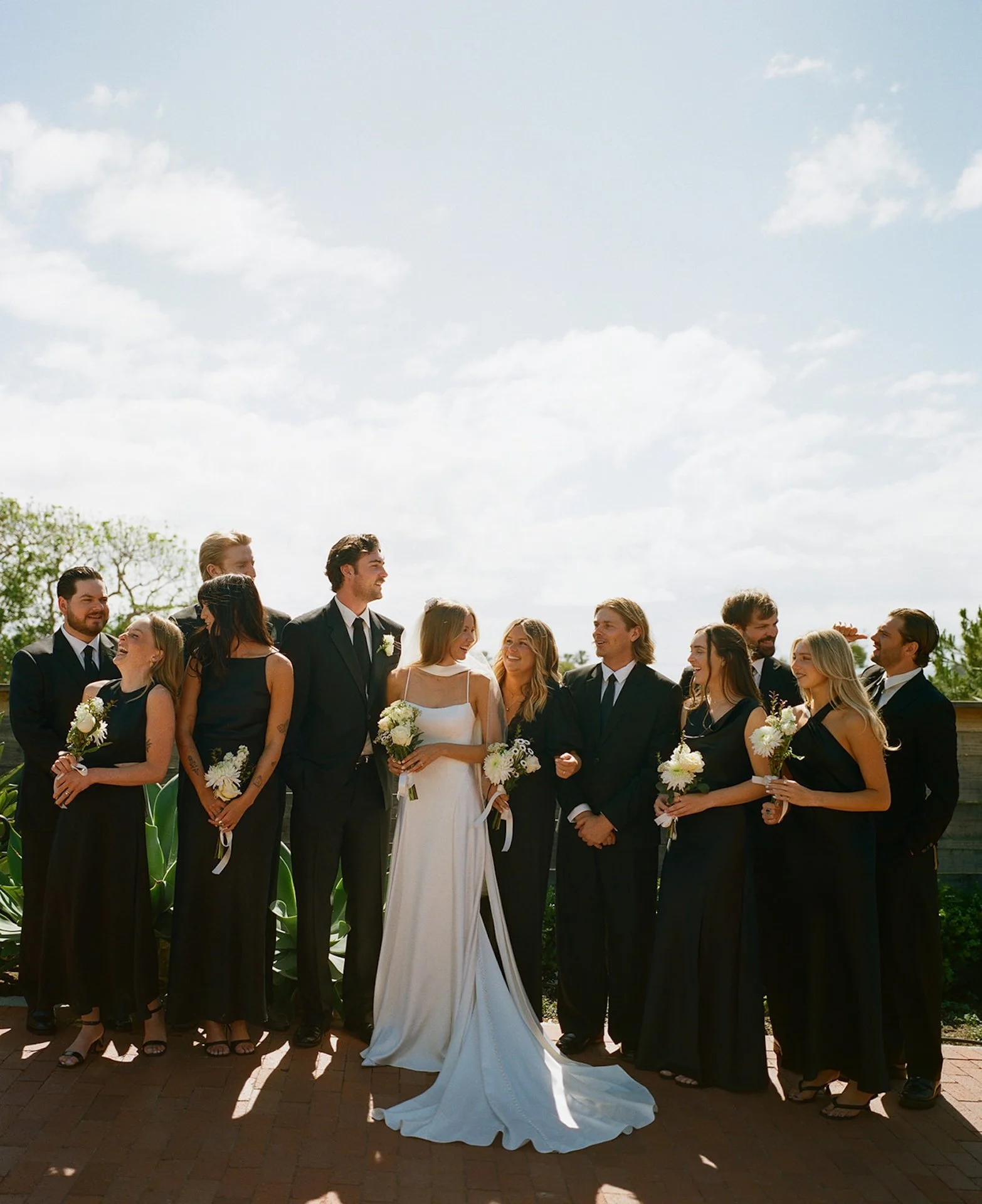 Wedding party standing together in black attire with the bride in a white dress during a sunny Laguna Beach wedding group portrait.