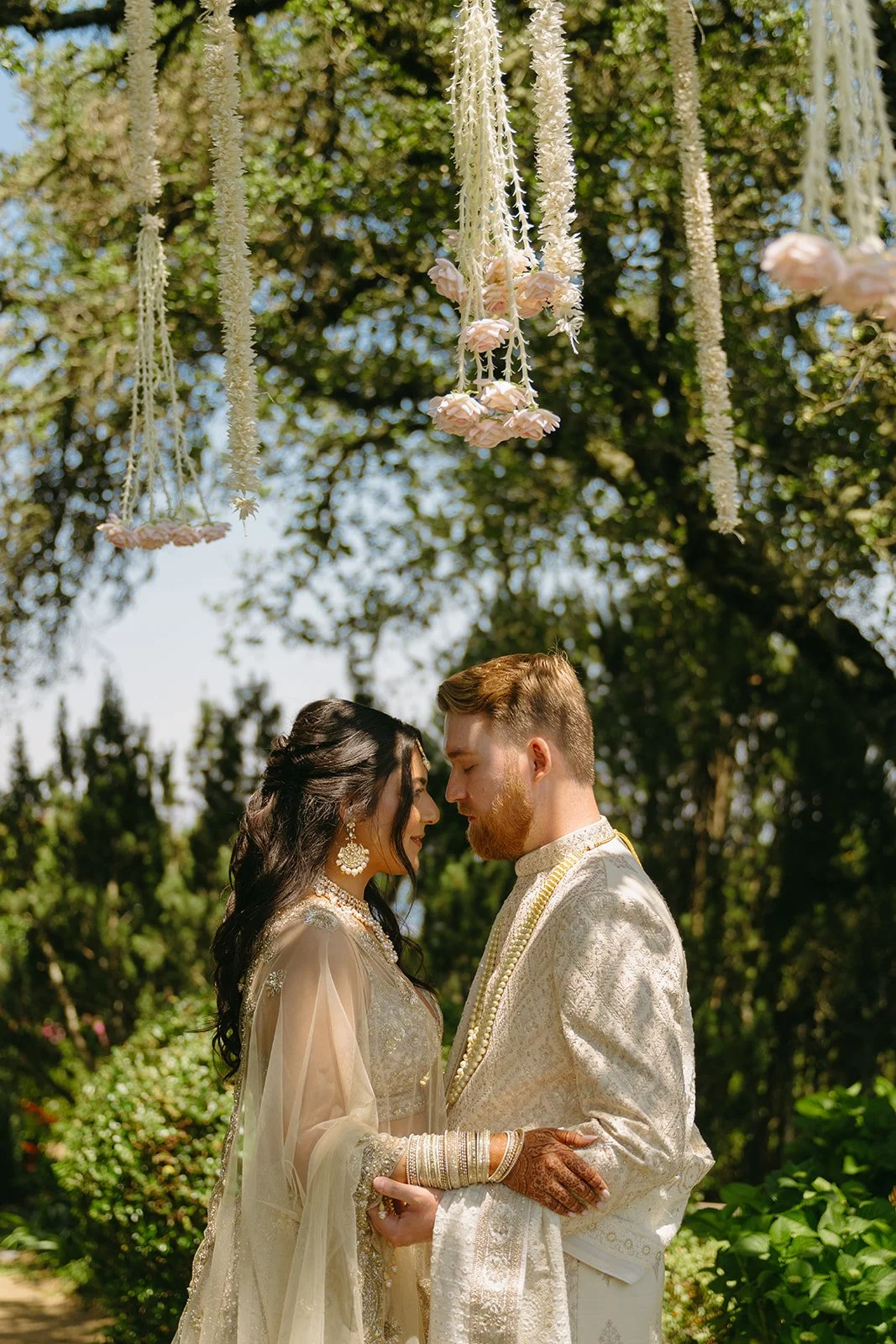 Couple in traditional attire standing close together beneath hanging florals during an intimate Indian wedding, photographed by a California wedding photographer.