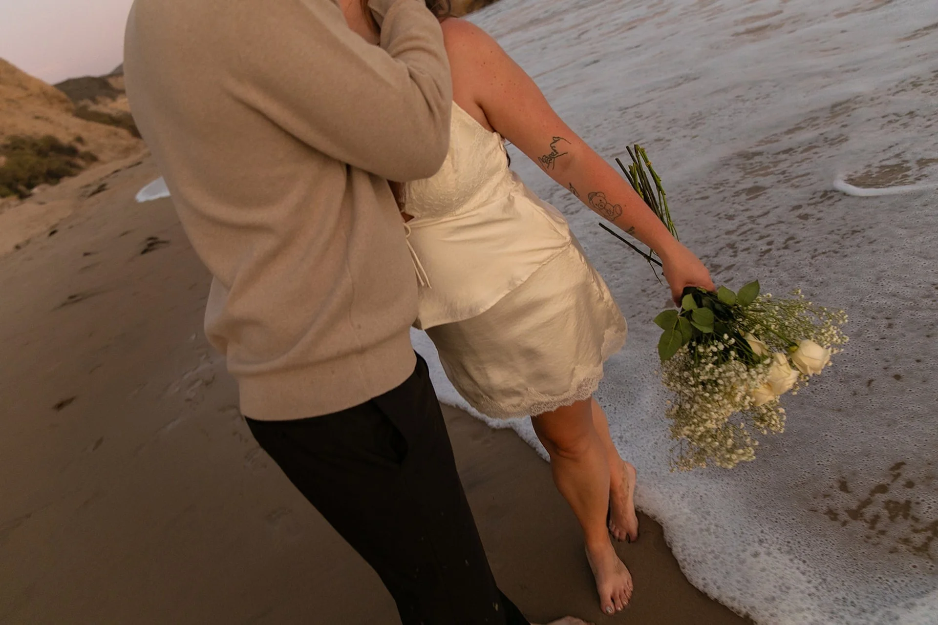 Beach Engagement Photo Ideas of a couple kissing while walking along the shoreline at sunset, with soft waves and pastel sky tones in the background.